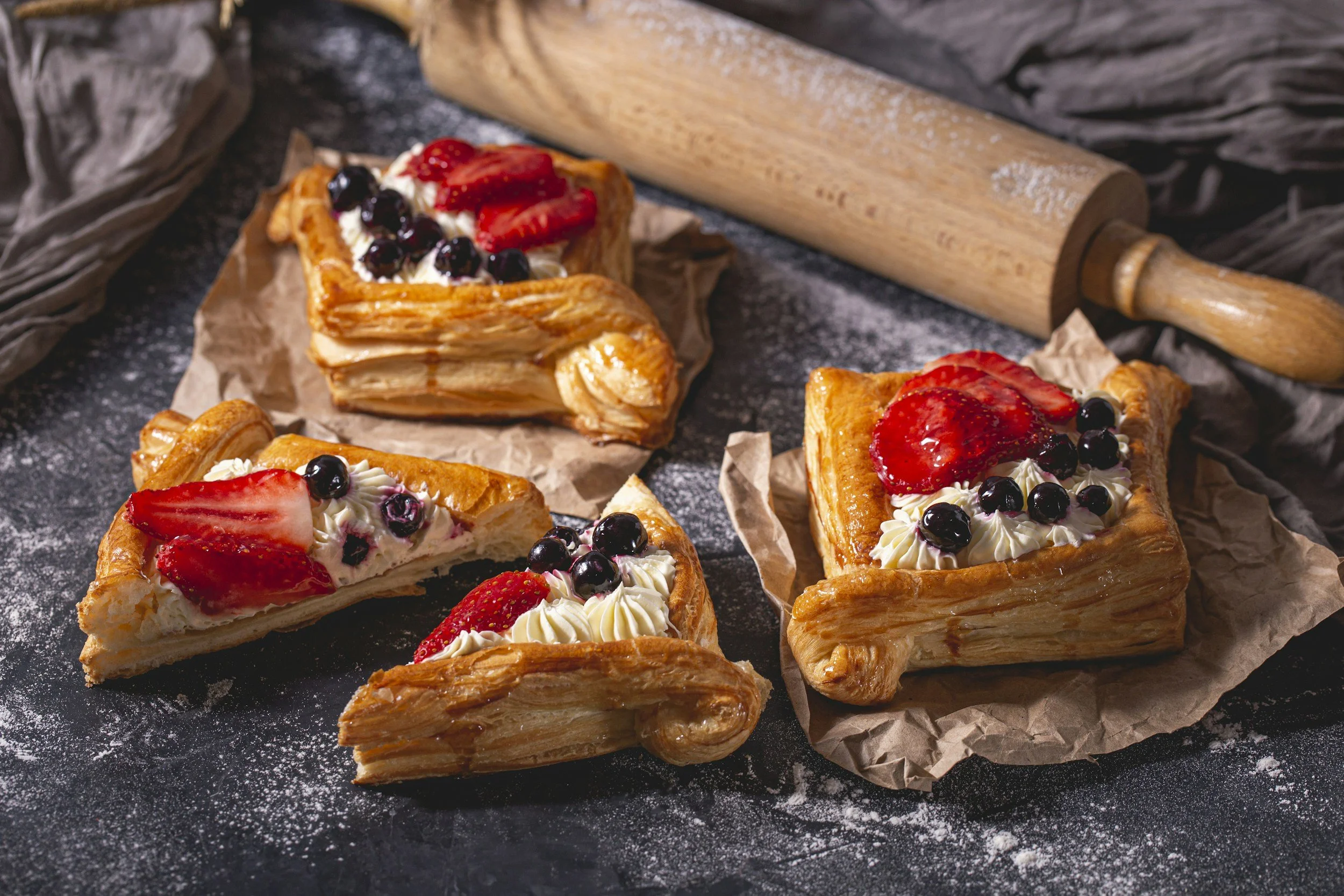 Assorted strawberry and blueberry Danish pastries on a dark surface with a pizza roller nearby.