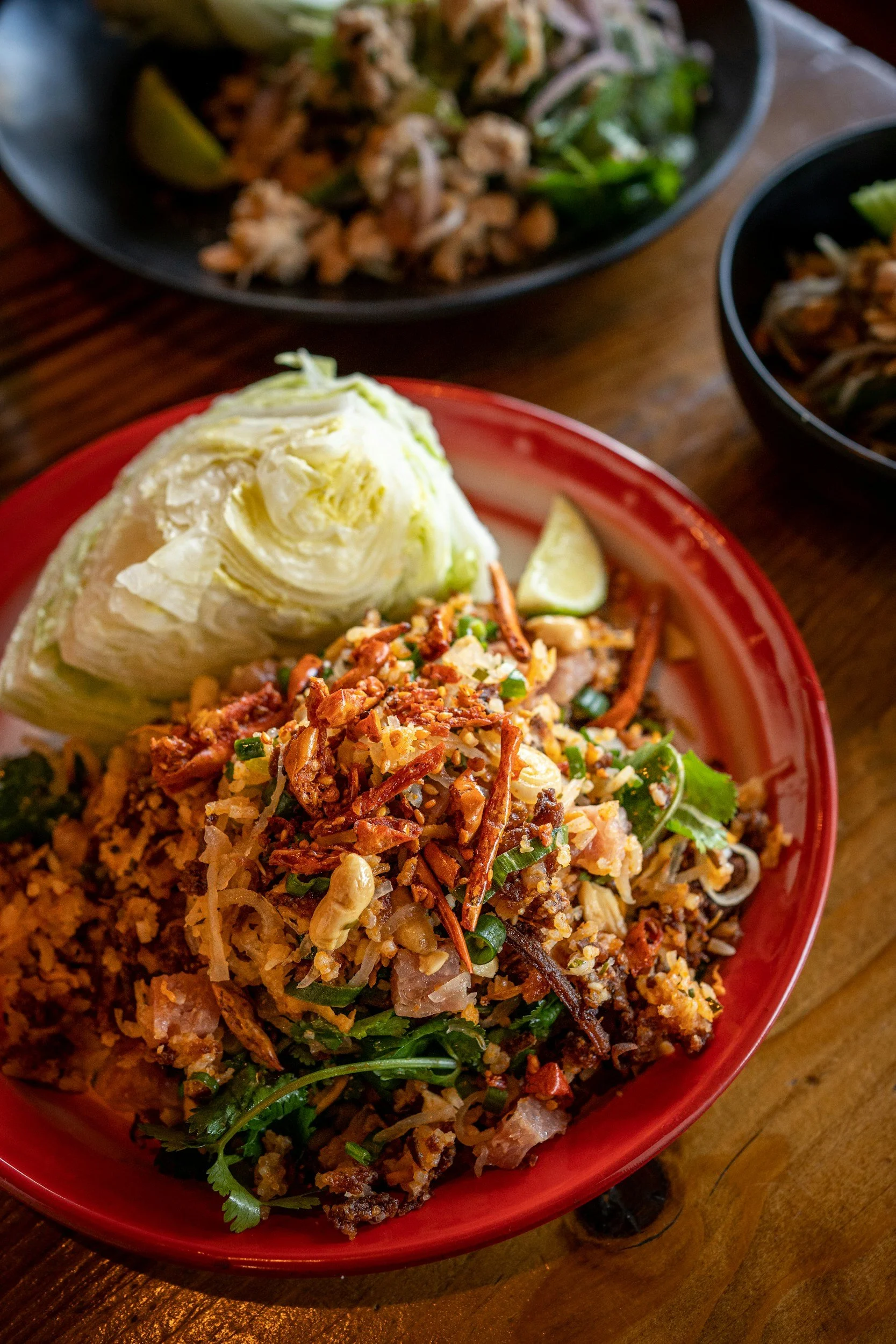 A bowl of fried rice topped with fried shallots and garnished with cilantro, with lime wedges on the side. In the background, there are two additional bowls of food.