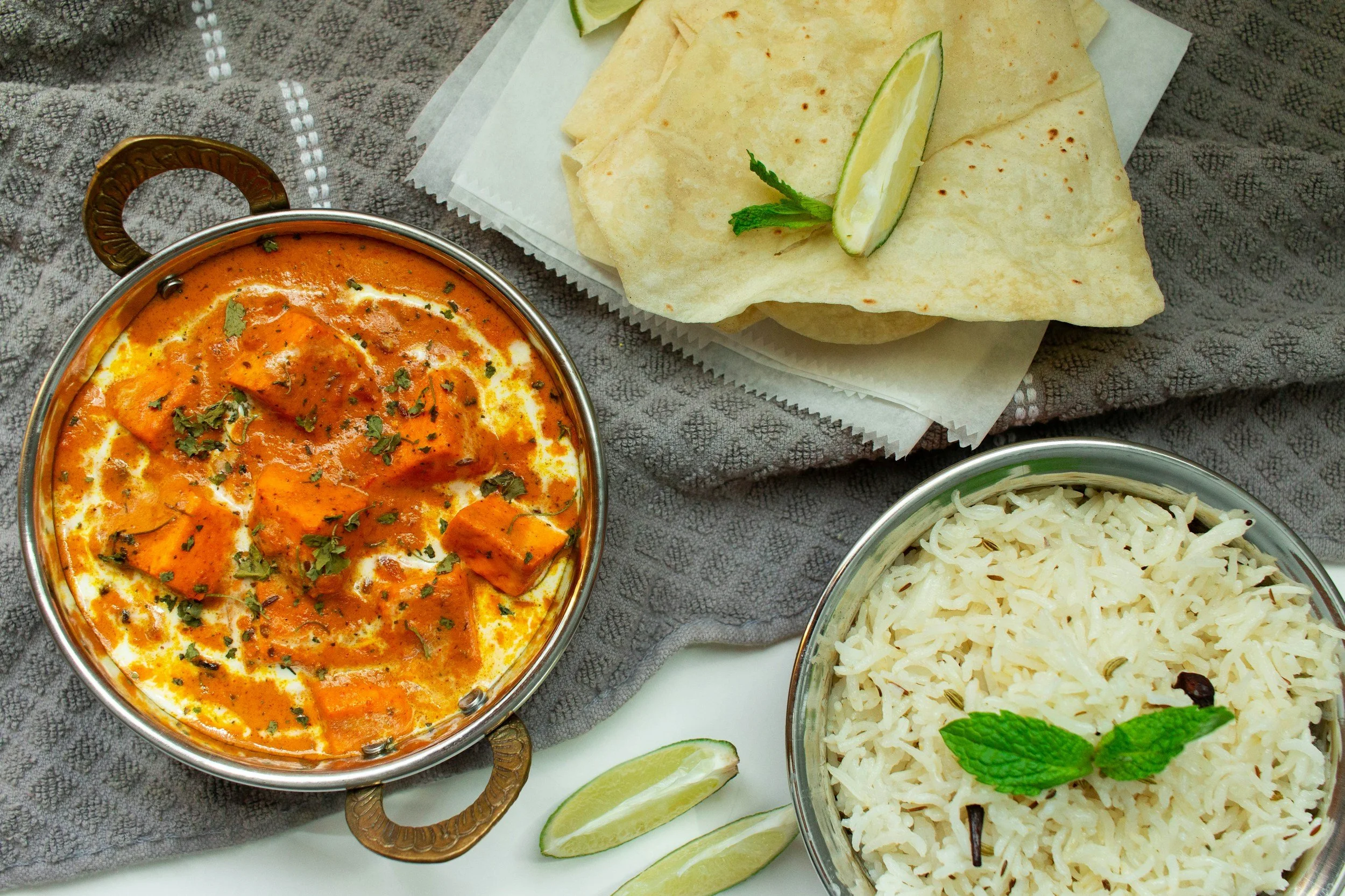 A plate of Indian curry with chunks of chicken in a spicy orange sauce, served with white rice garnished with mint leaves, and accompanied by folded naan bread with lime wedges on a gray patterned cloth.