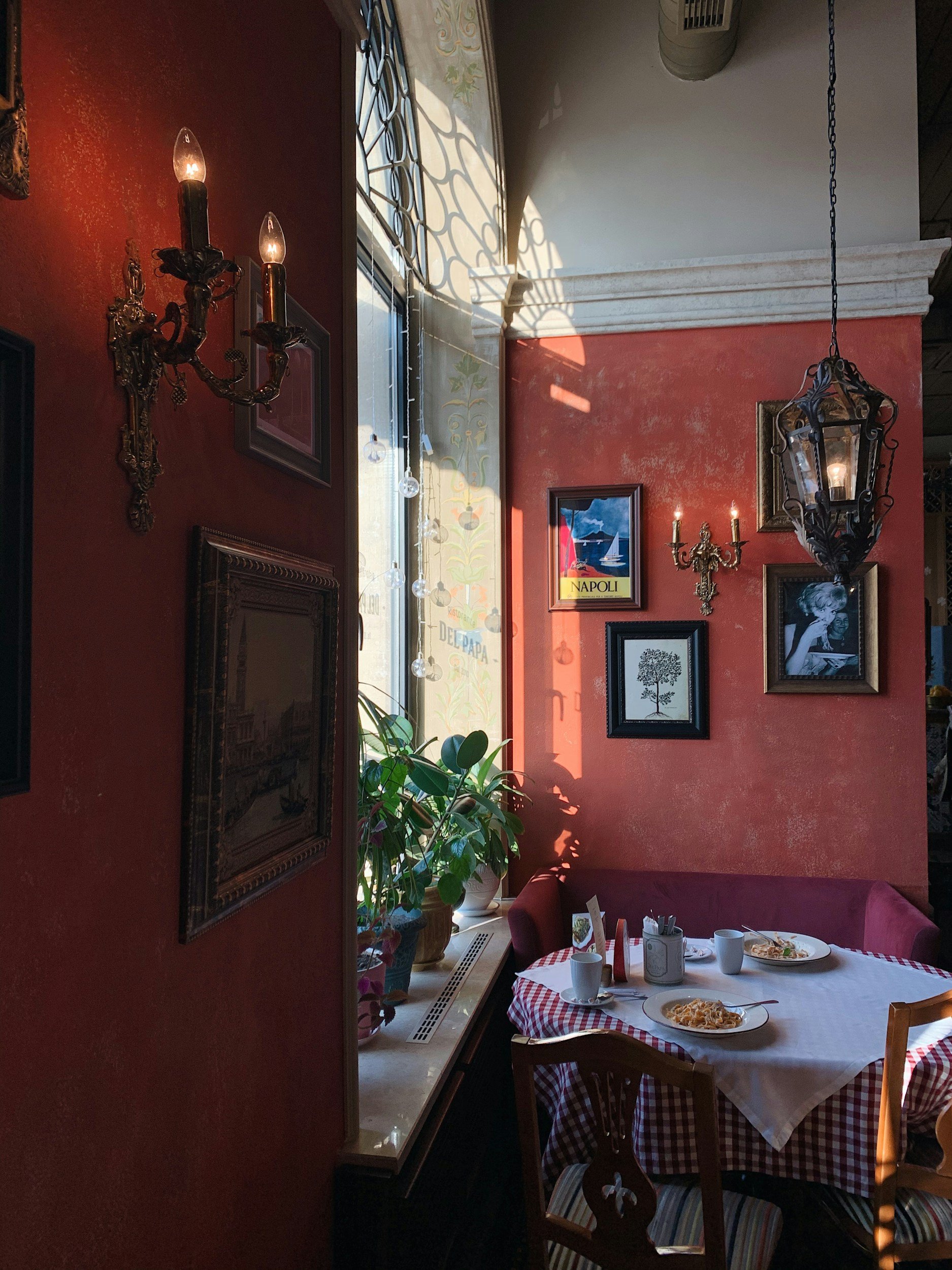 A cozy dining area with a table covered with a red and white checkered tablecloth, set with plates of food, mugs, and utensils. The room has red textured walls with framed artwork, black sconces, and a large hanging lantern. Sunlight streams through a window, casting shadows on the wall.