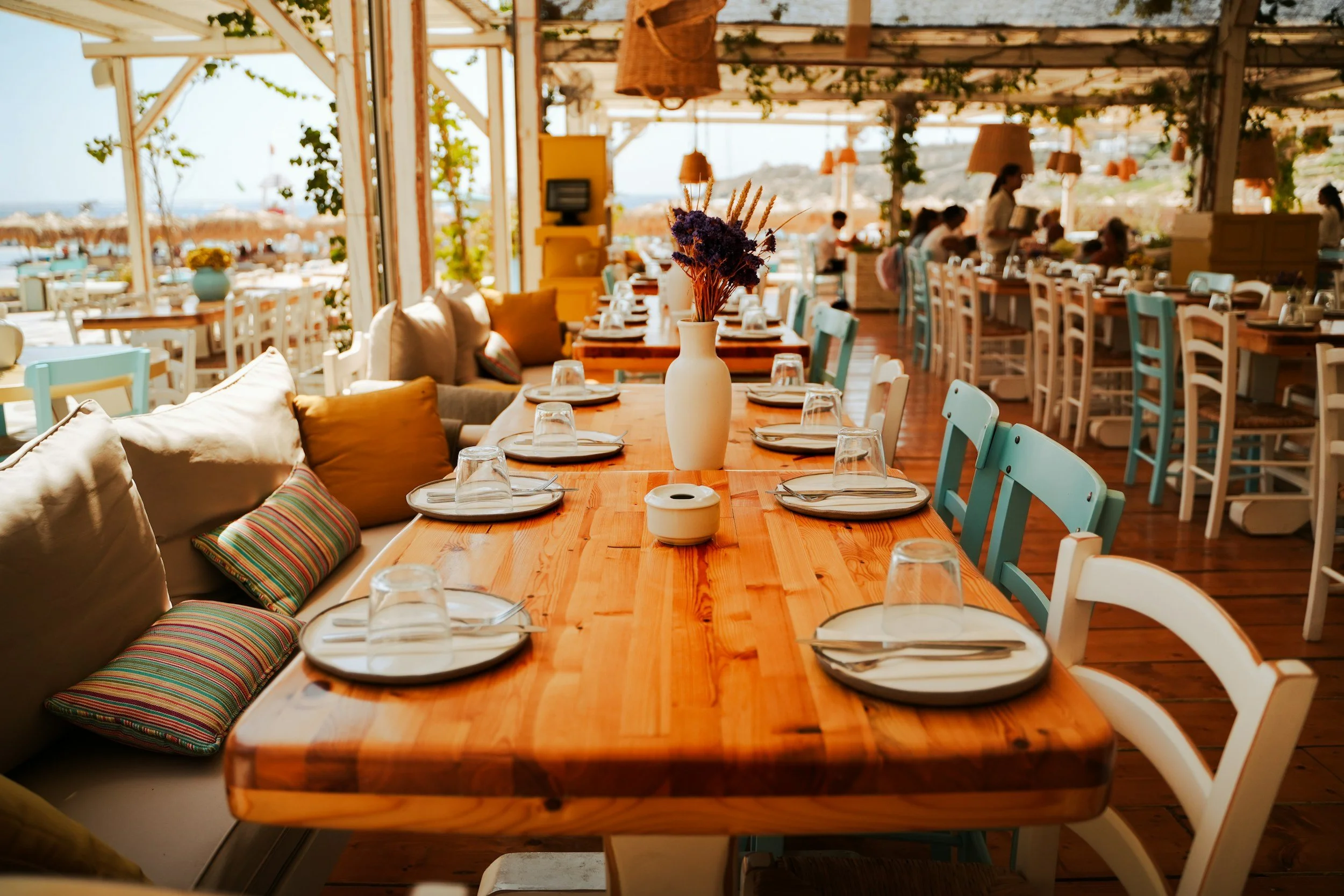 A bright, airy restaurant with a wooden dining table set with placemats, glasses, and a white vase with purple flowers. The restaurant has colorful chairs and cushions, with sunlight coming through large windows showing an outdoor view of a beach with straw umbrellas.