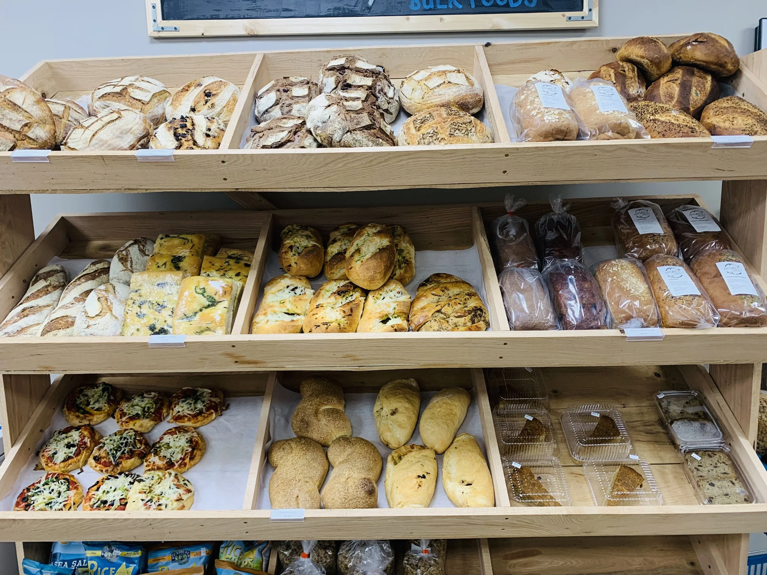 Wooden display shelves filled with various types of freshly baked bread and pastries, including loaves, rolls, and flatbreads at a bakery.