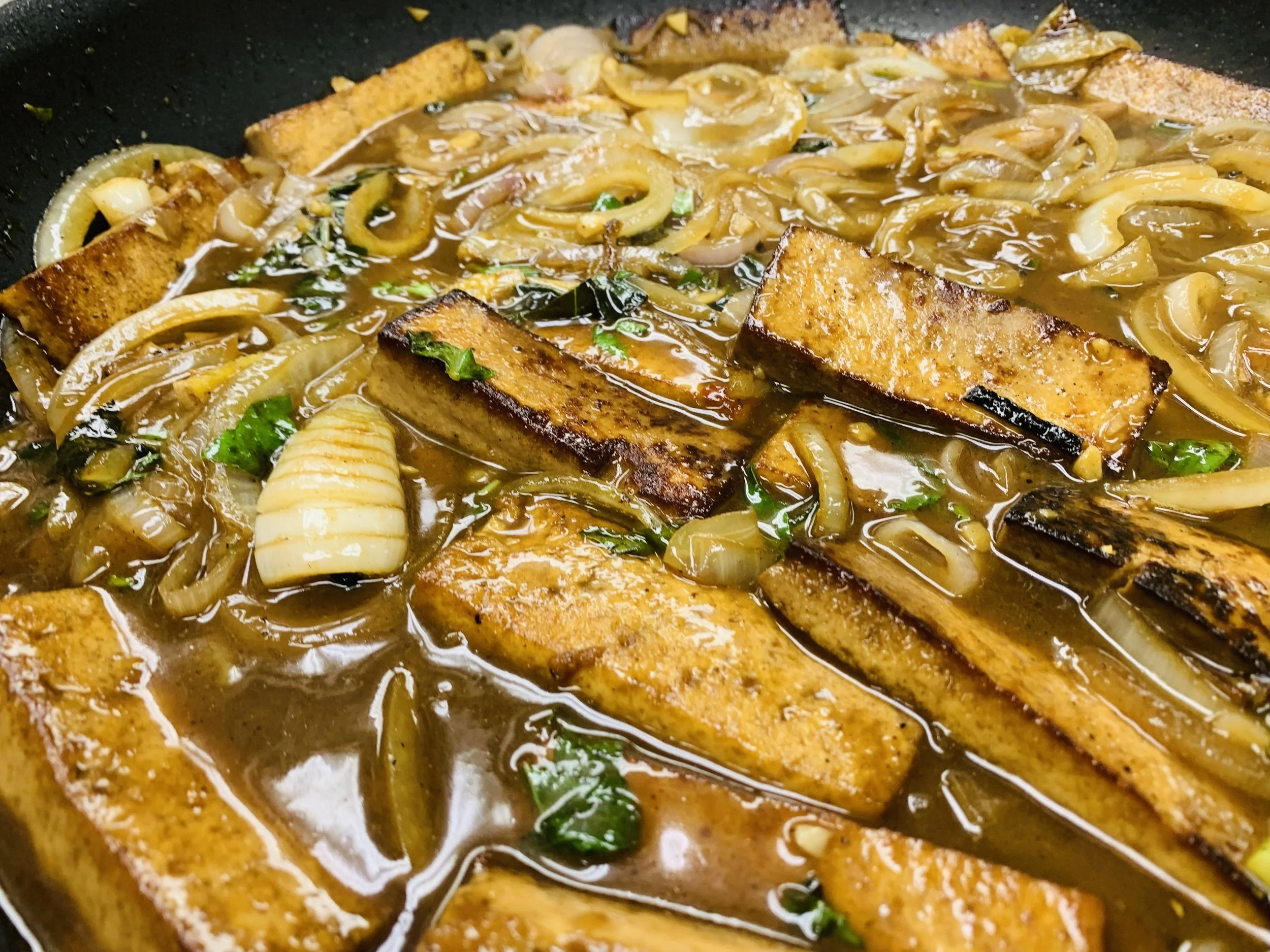 Close-up of a skillet with cooked fish fillets in a sauce with onions, herbs, and vegetables.