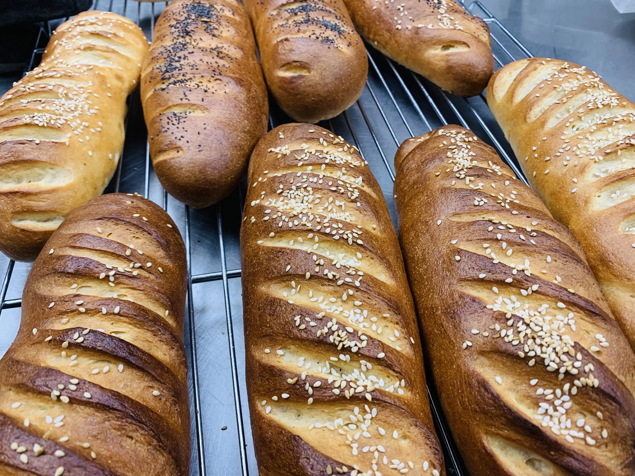Several freshly baked baguettes on a wire rack, some topped with sesame seeds and others with poppy seeds.