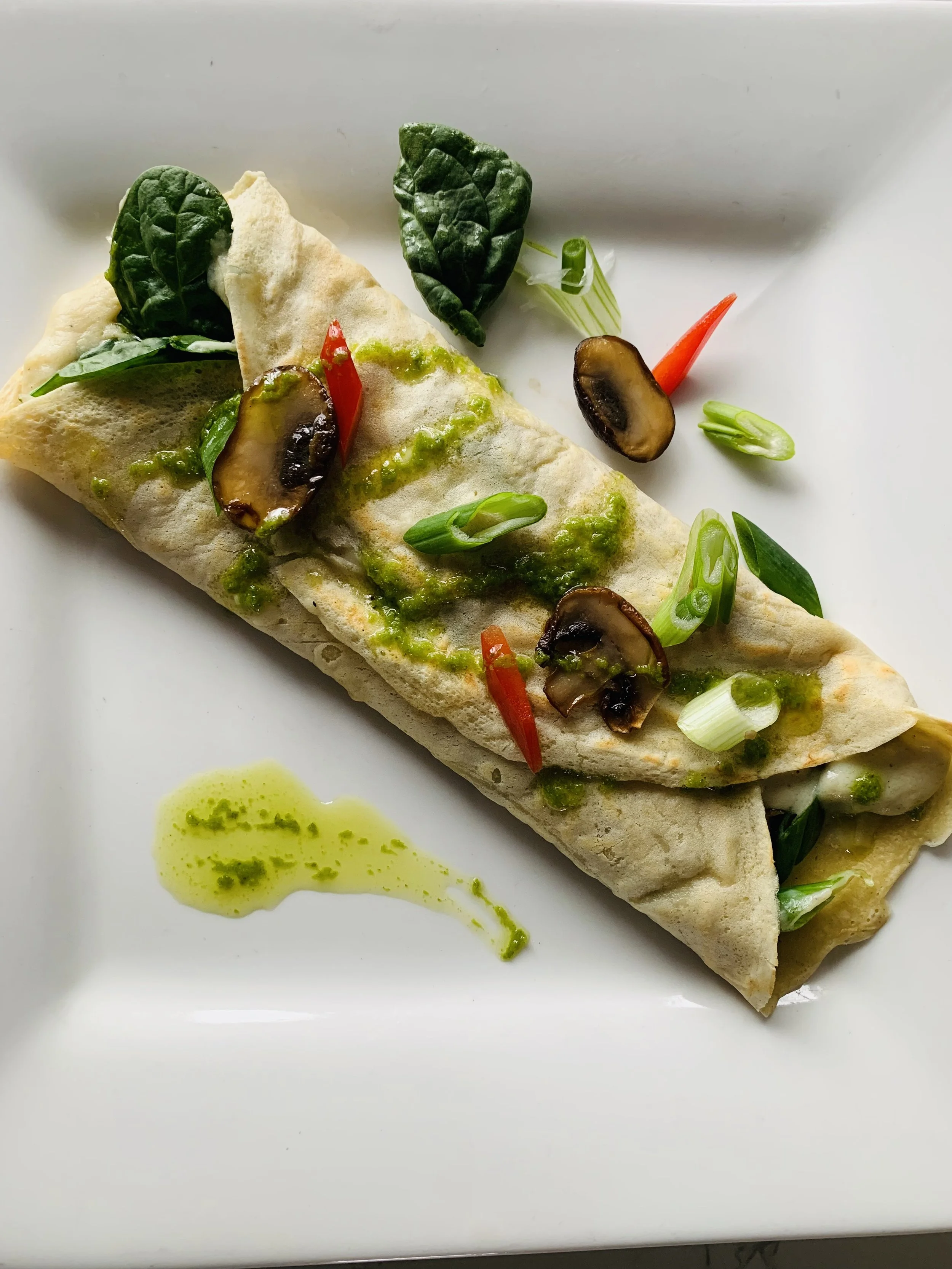 Close-up of a rolled wrap or burrito garnished with green sauce, sliced mushrooms, chopped green onions, and small red peppers on a white plate.
