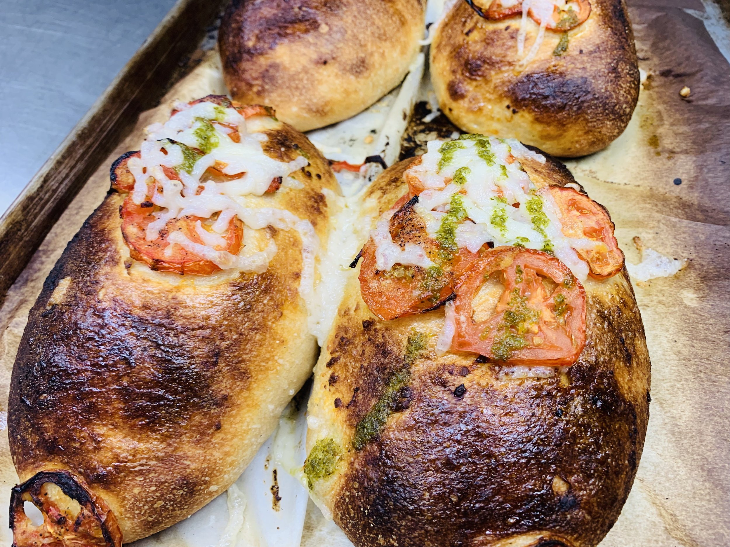 Close-up of three baked bread rolls topped with melted cheese, sliced tomatoes, and green sauce on a wooden tray.