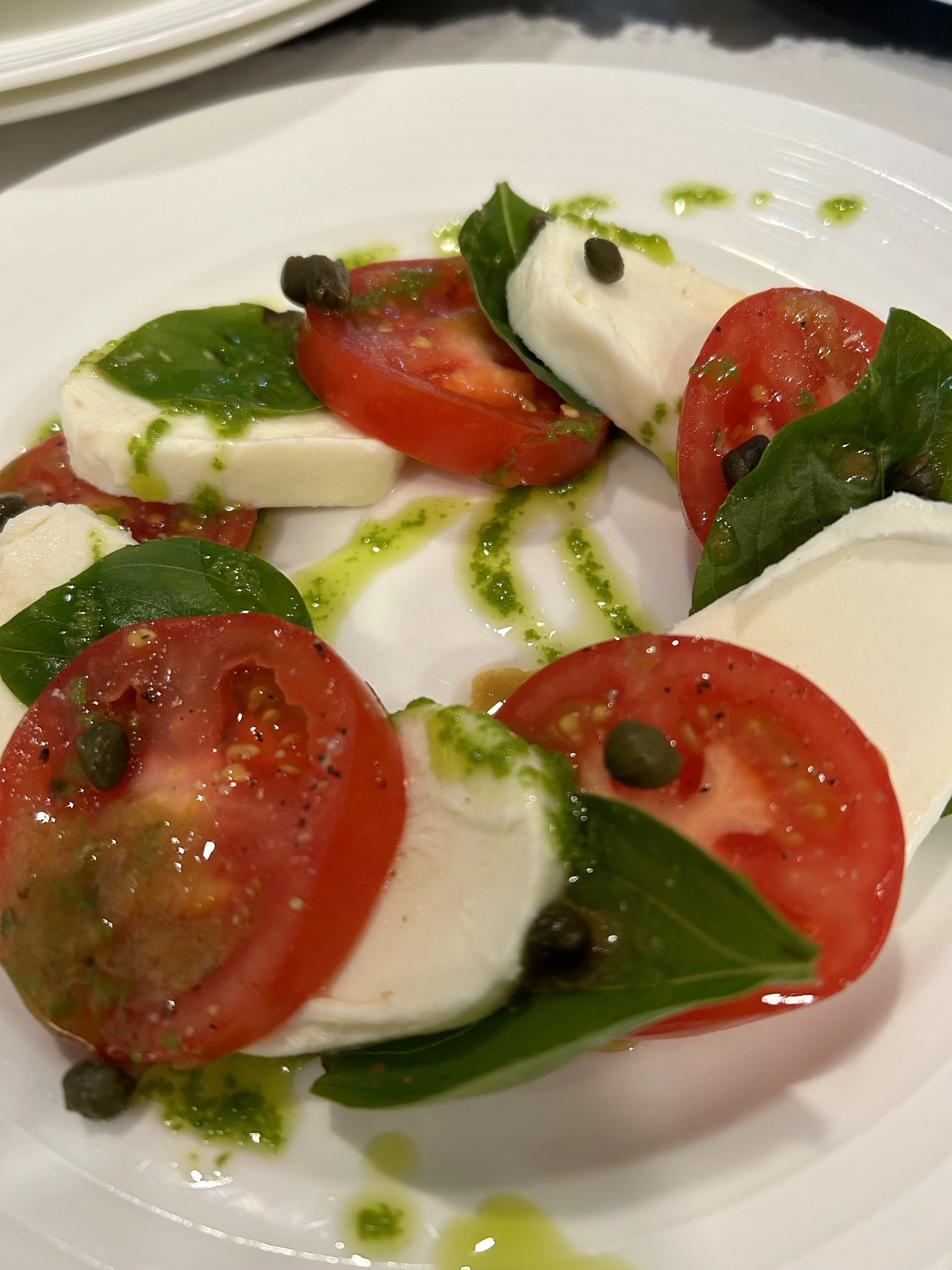 A Caprese salad with slices of tomato, fresh mozzarella, basil leaves, black pepper, and olive oil on a white plate.