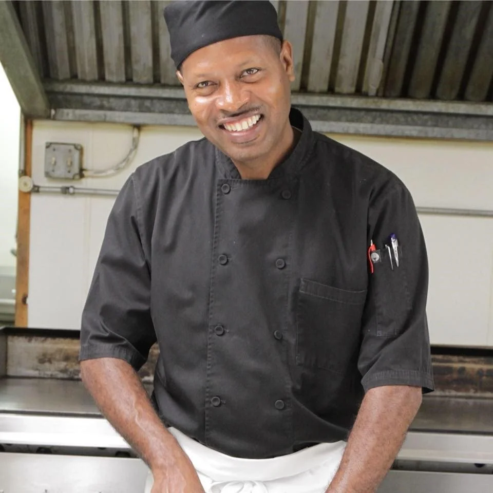 A smiling male chef in a black chef uniform and hat, in a kitchen.