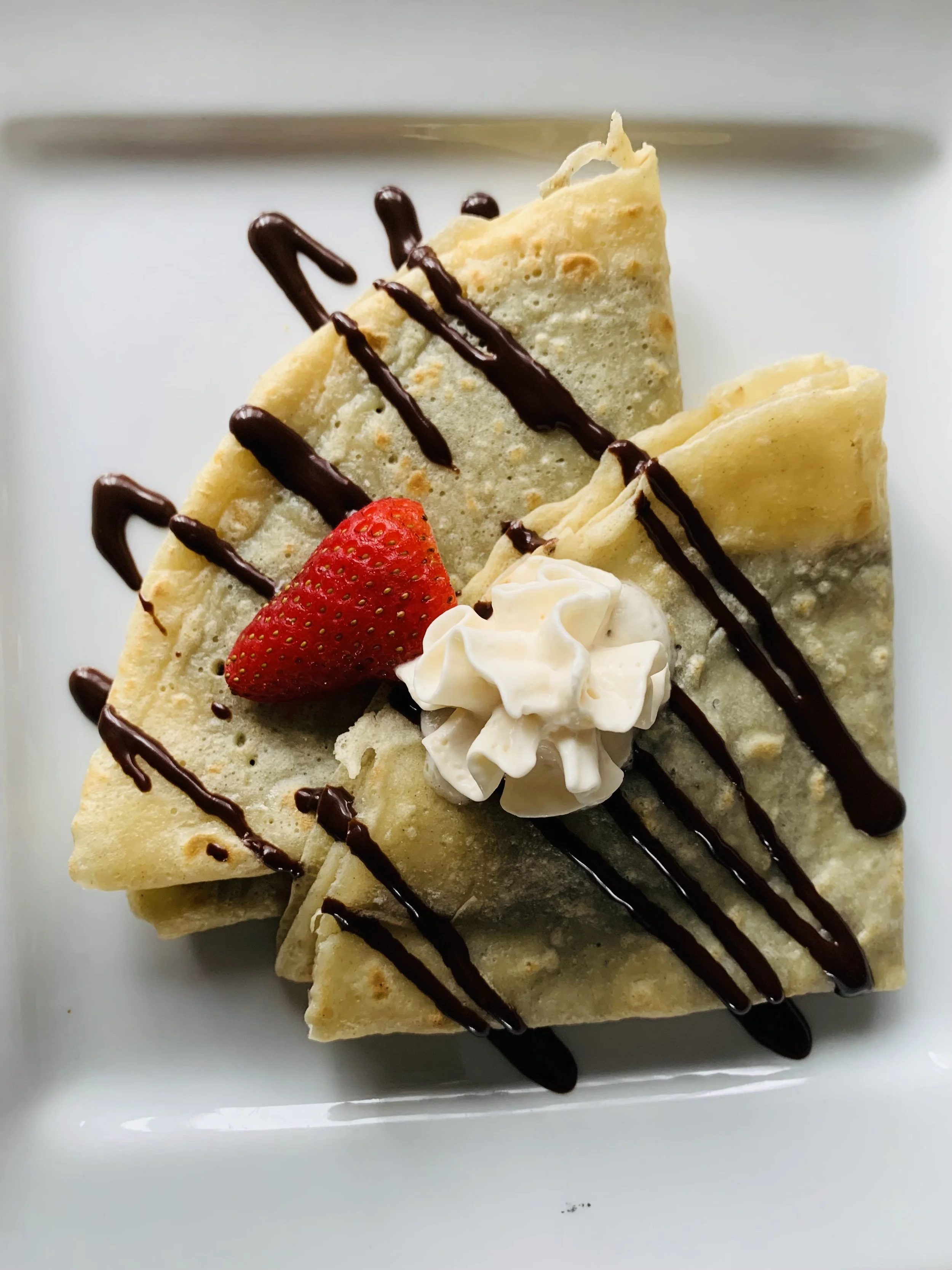 Close-up of folded crepes topped with a strawberry, whipped cream, and drizzled with chocolate sauce on a white plate.
