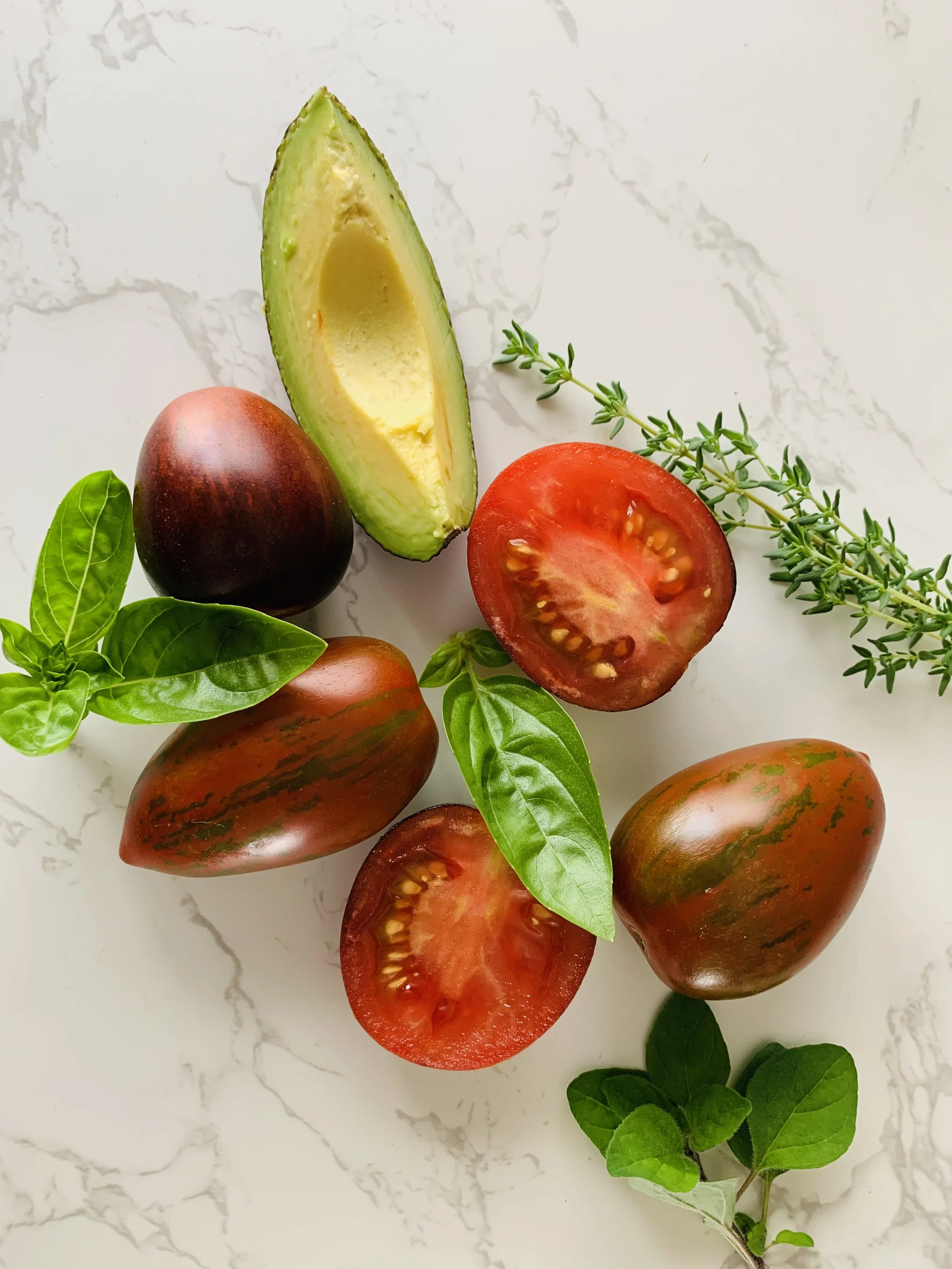 A collection of fresh vegetables and herbs including a halved tomato, a halved avocado, a whole plum tomato, a whole heirloom tomato, green basil leaves, and sprigs of thyme on a white marble surface.