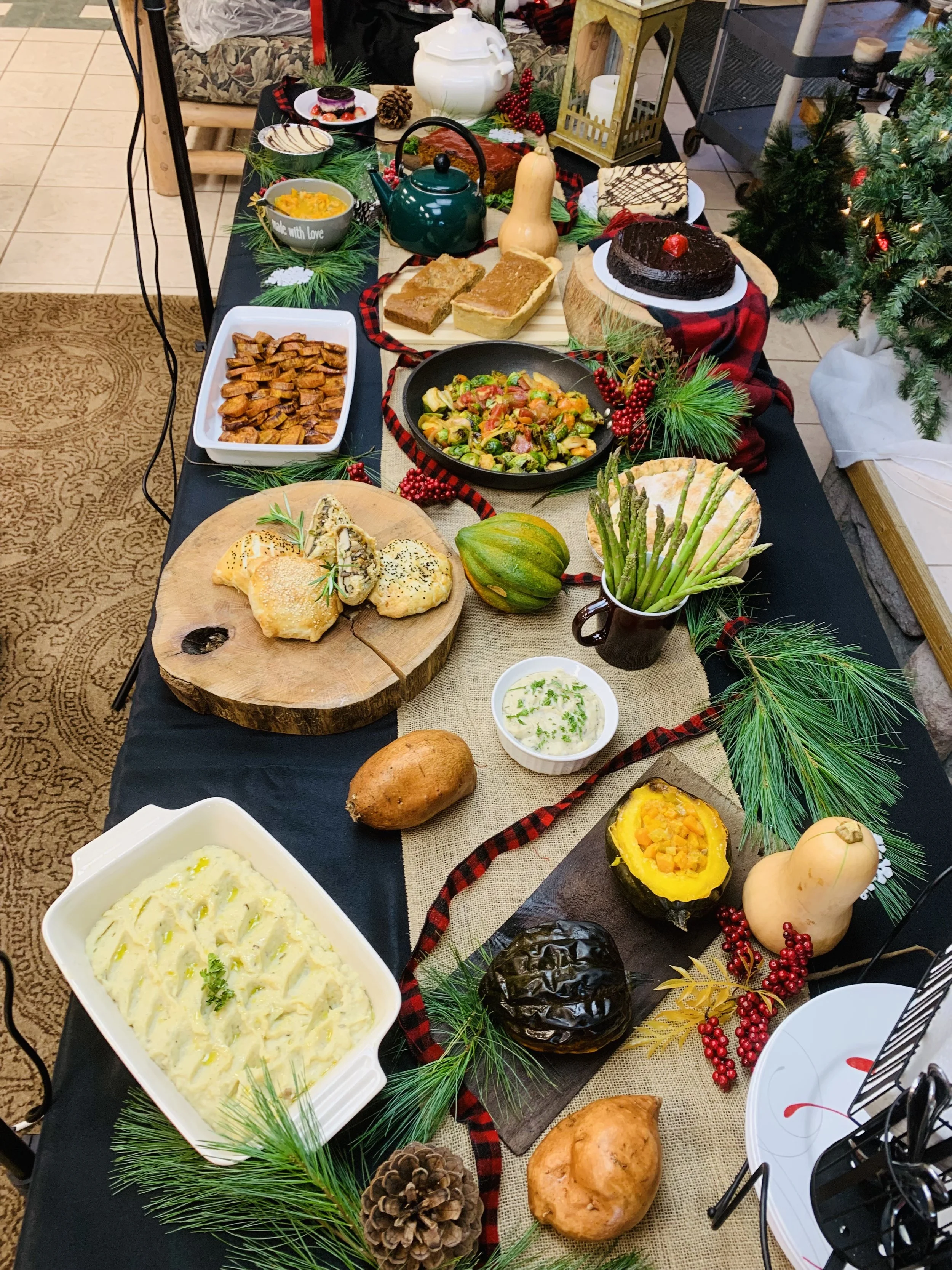 A festive holiday table with various dishes including baked bread, a casserole, mashed potatoes, soup, roasted vegetables, and desserts. The table is decorated with pine branches, pinecones, berries, and a red and black plaid ribbon.