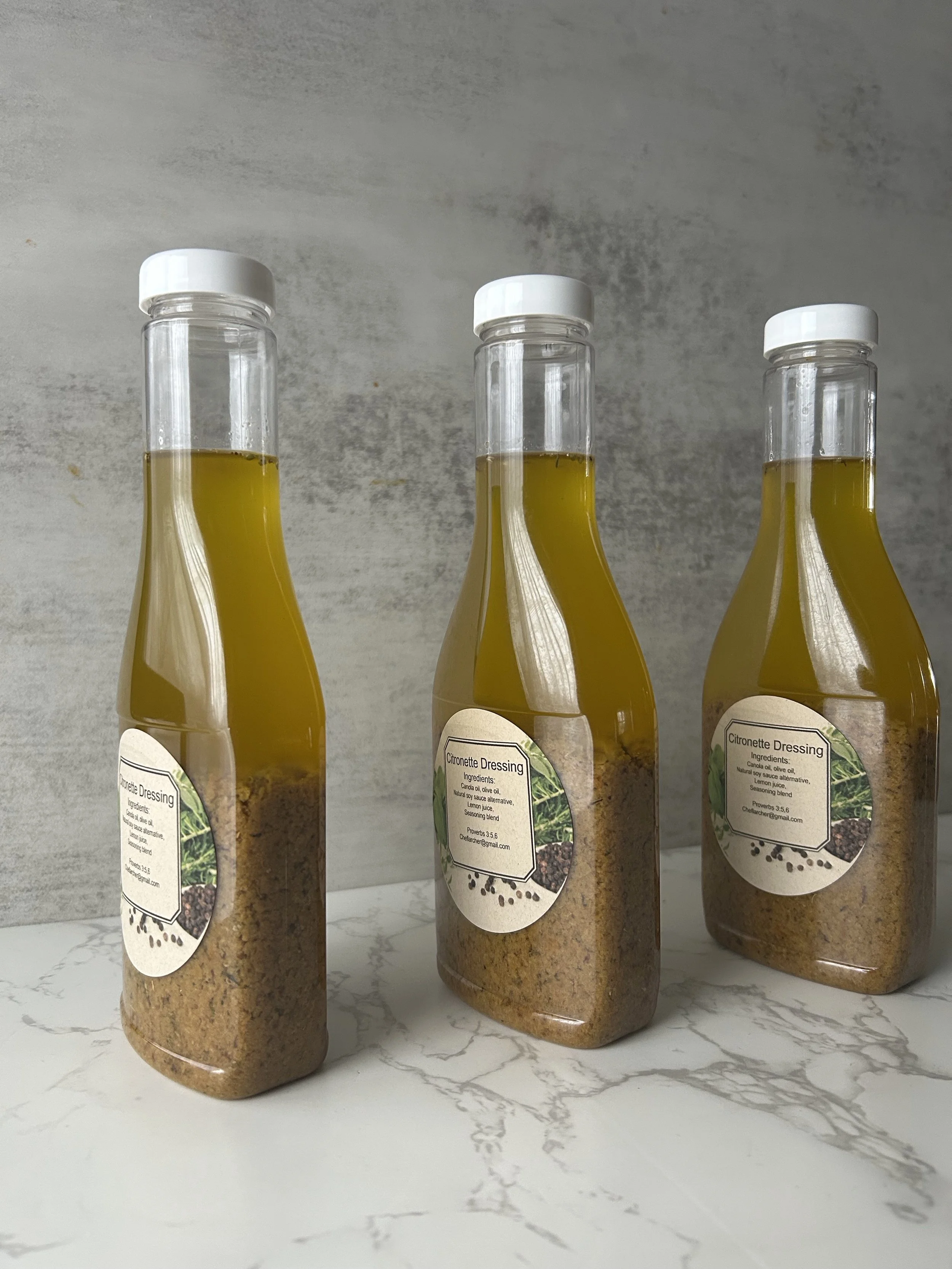 Three bottles of citronette dressing with yellow liquid, standing upright on a white marble surface. Each bottle has a white cap and a label with ingredients and contact information.