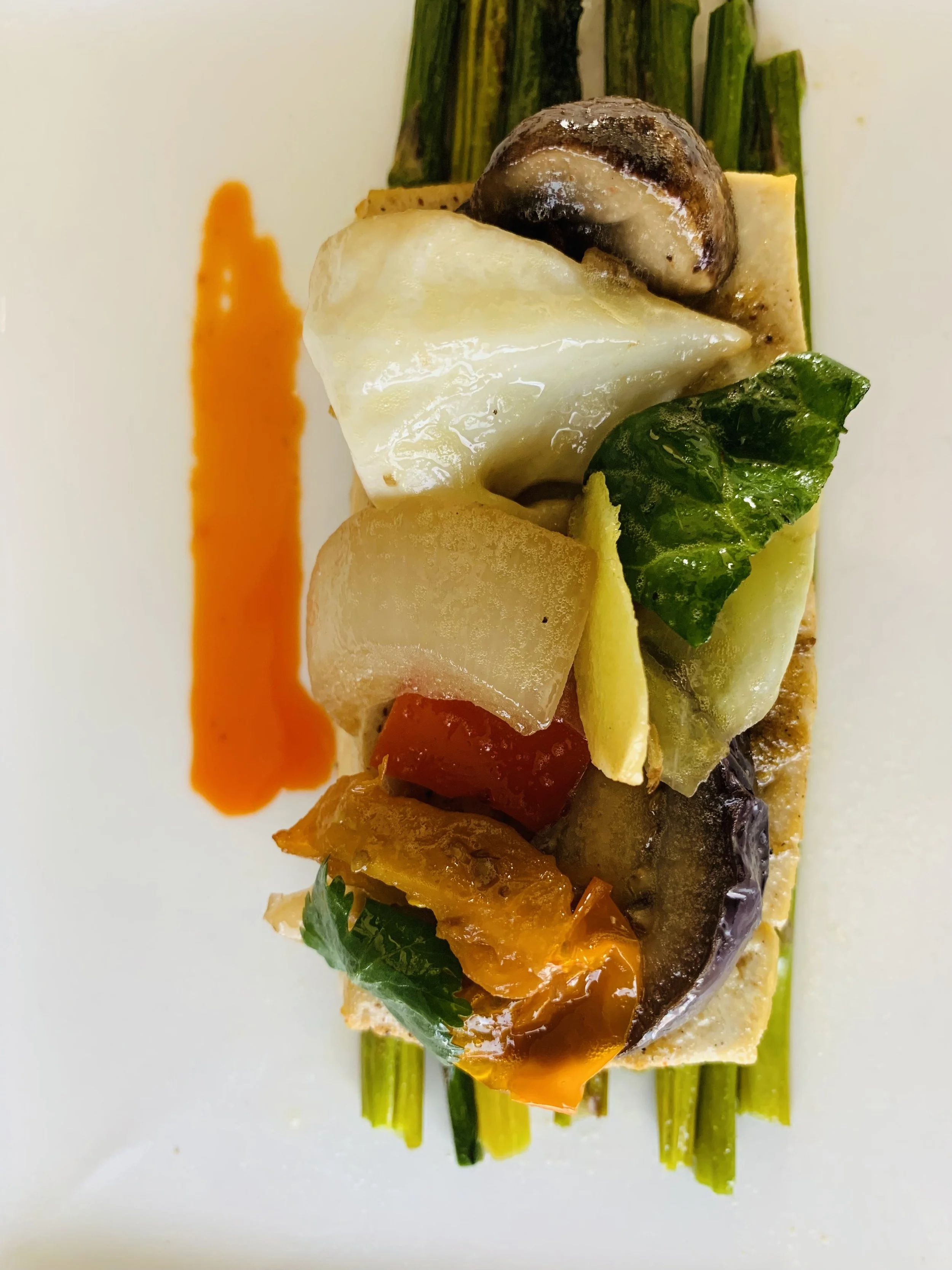 A plated appetizer featuring green asparagus spears, a slice of cheese topped with a cooked mushroom cap, roasted vegetables including a cherry tomato and peppers, with a streak of red sauce on the side.