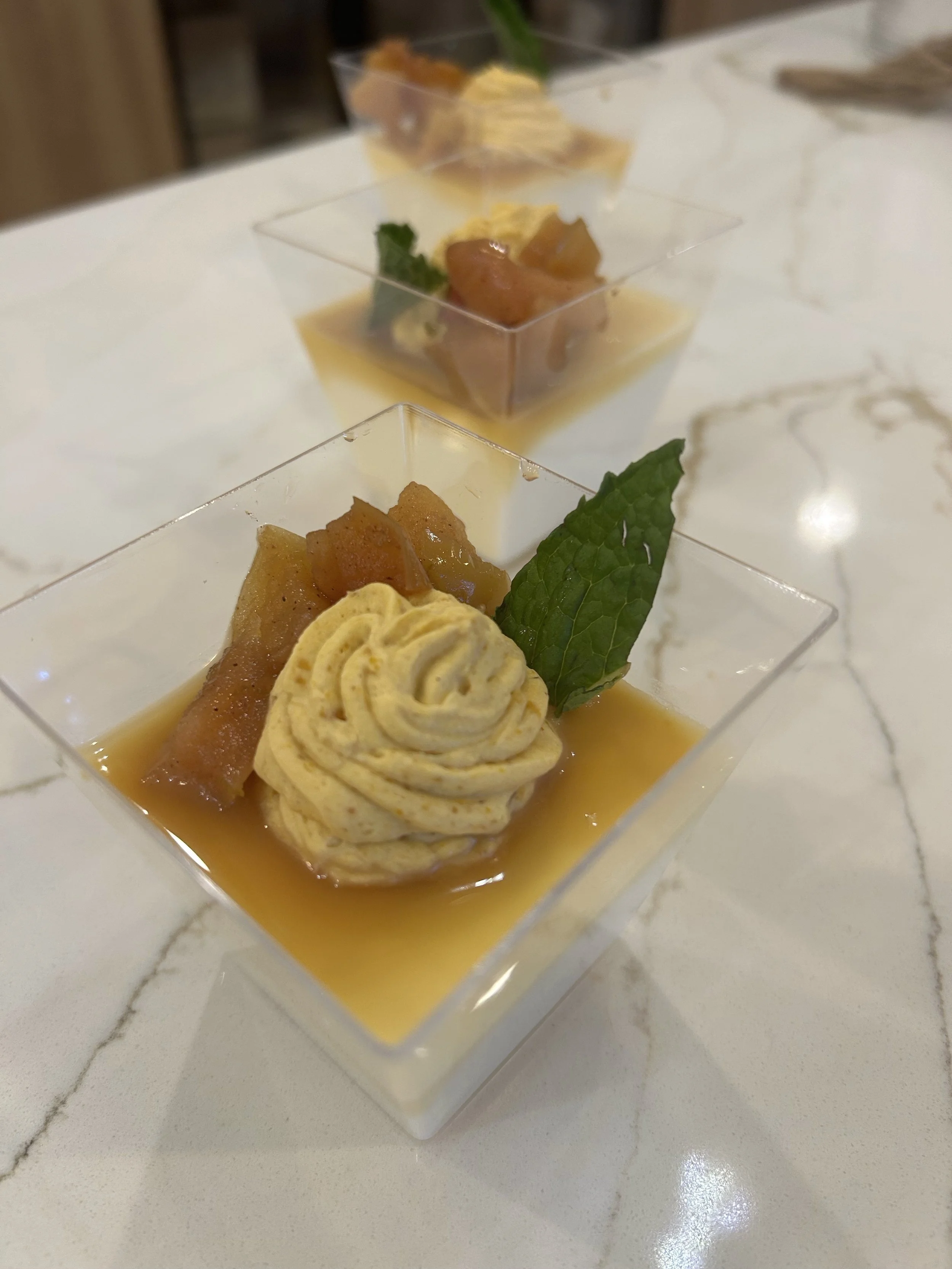 Dessert in a clear square container with a garnish of mint leaf, caramelized fruit, and rolled pastry with sauce.