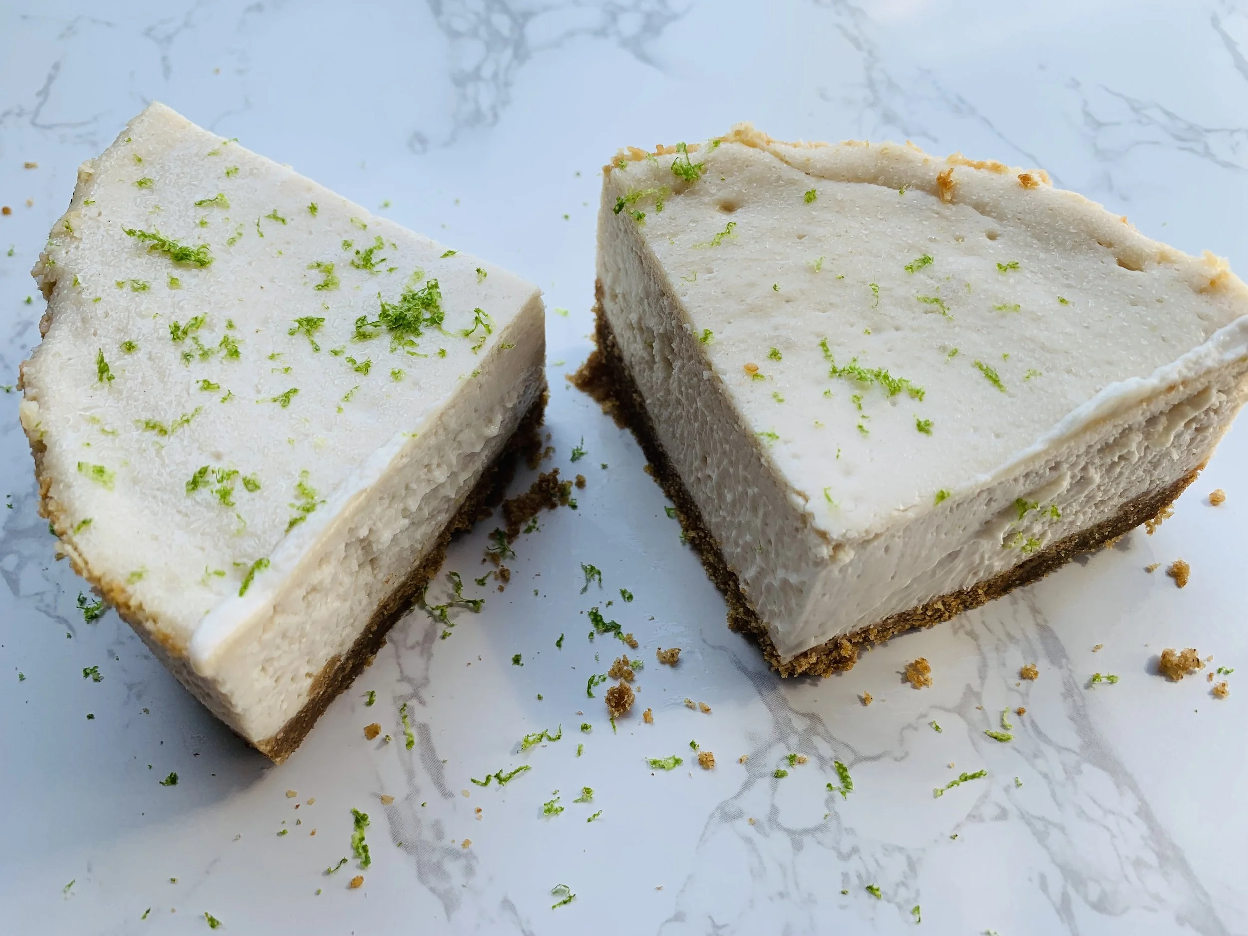 Slices of creamy lime-flavored cheesecake with a graham cracker crust, garnished with lime zest, on a white marble surface with crumbs around.
