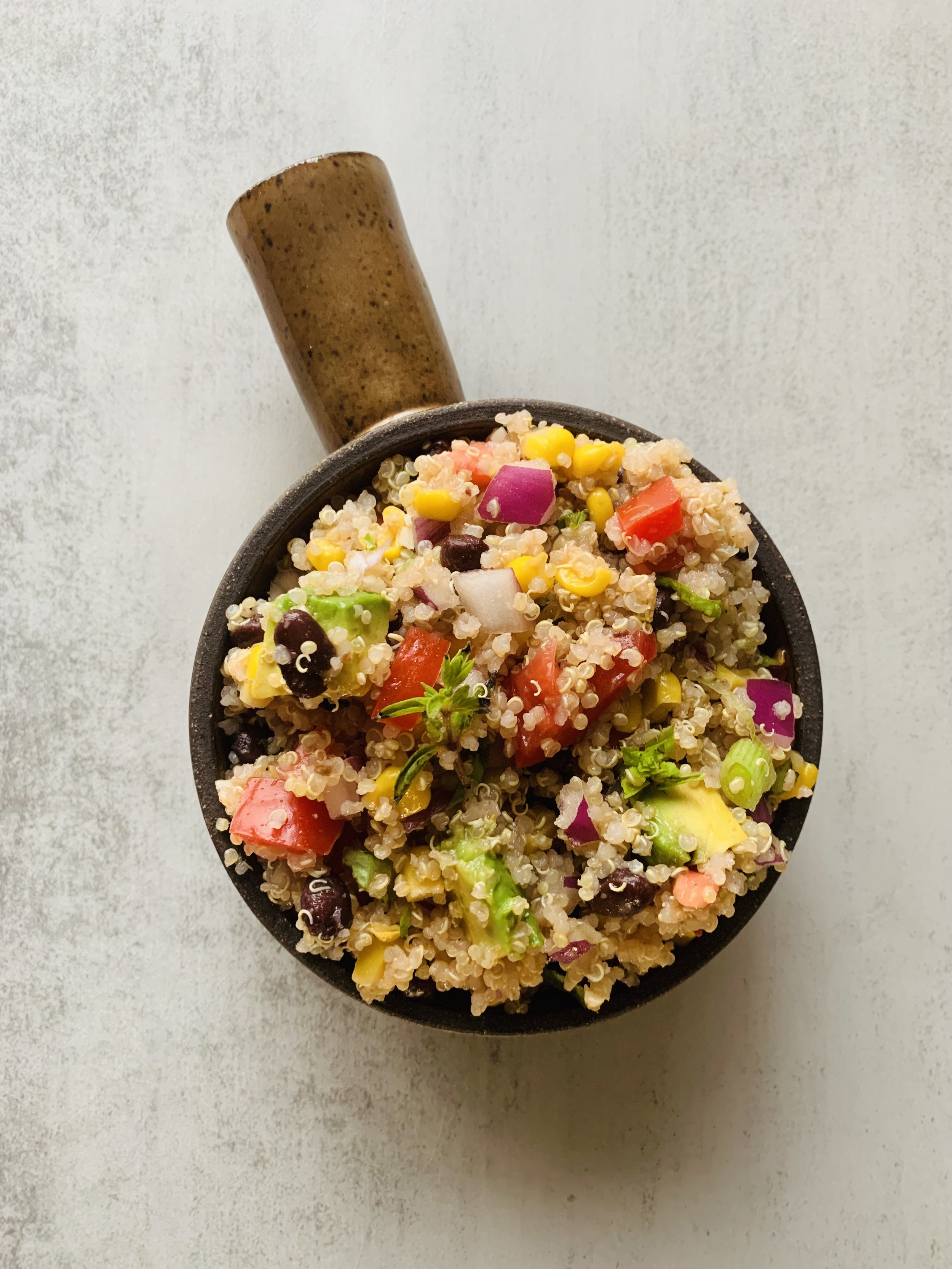 A bowl of quinoa salad containing diced red onions, cherry tomatoes, black beans, yellow corn, chopped parsley, and green onions, with a pepper grinder in the background.