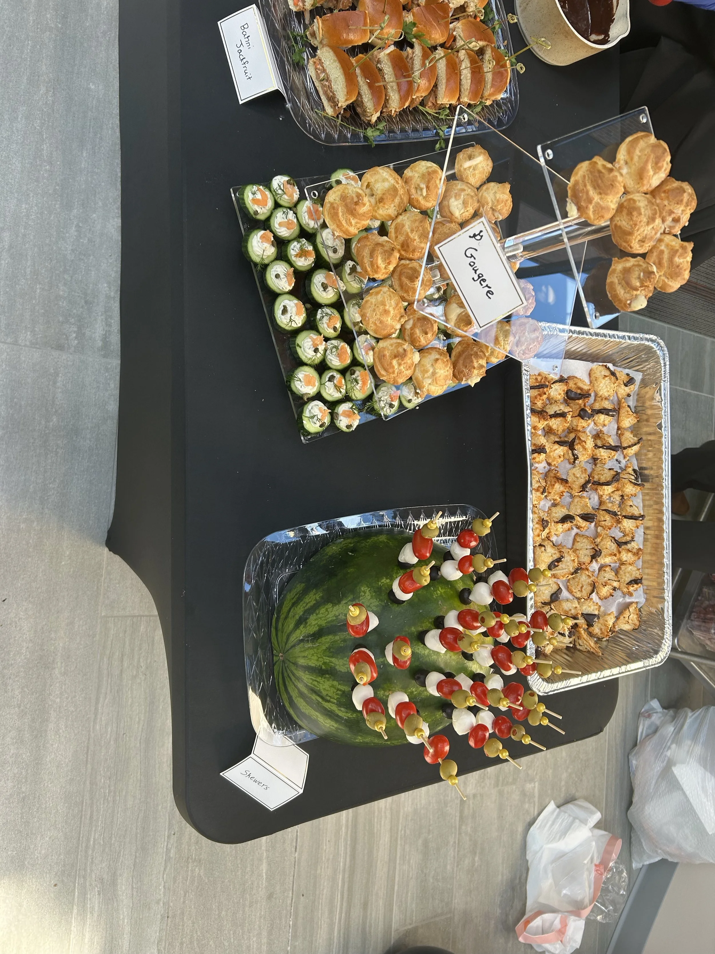 A table displaying various finger foods and snacks includes cucumber rolls with cream cheese and smoked salmon, cheese and meat skewers, small buns, and sliders. There are labels identifying the sections, such as 'Sowers', 'Mini Sandwiches', and 'Gou