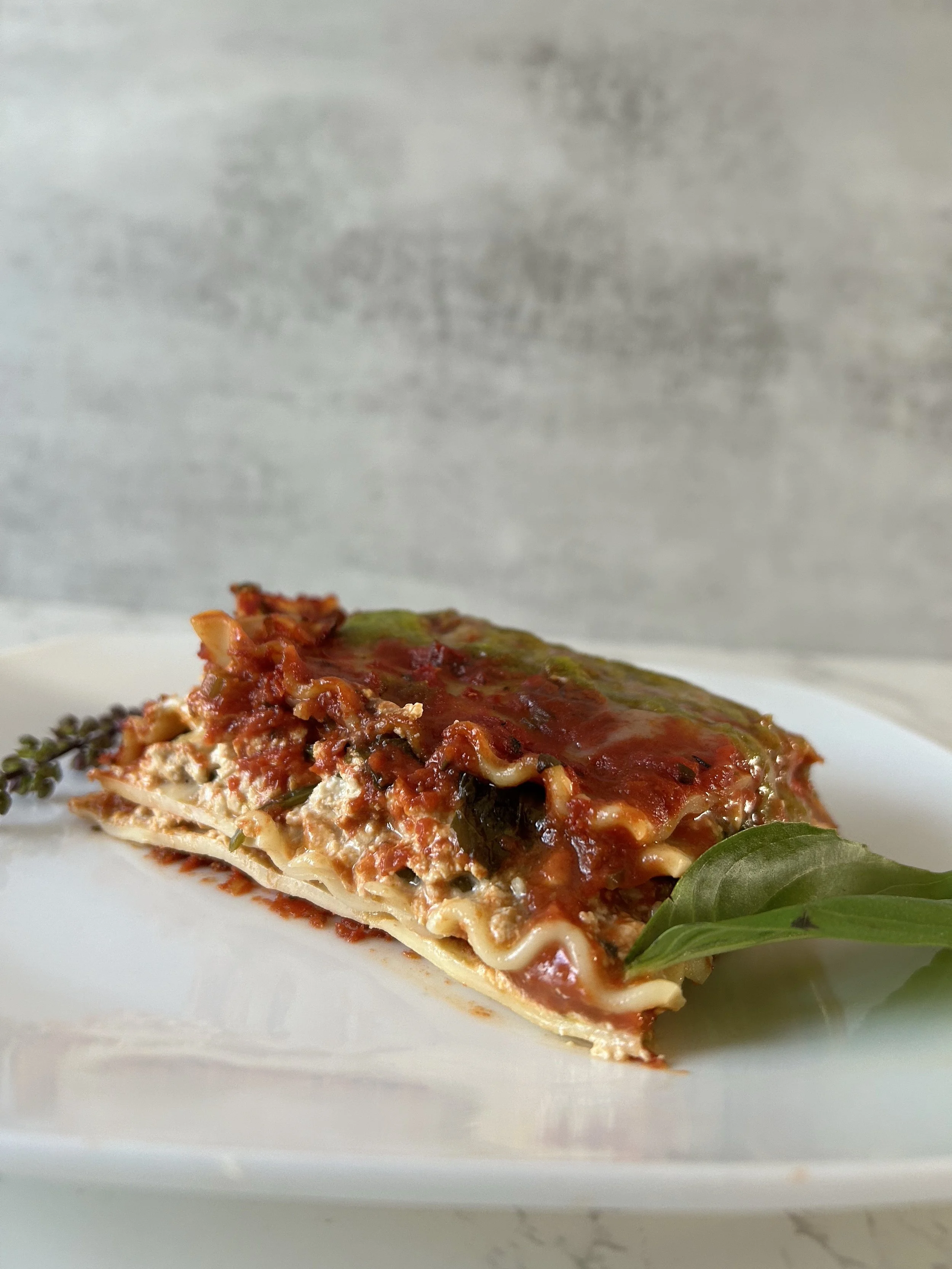 A slice of lasagna with layers of pasta, meat sauce, cheese, and tomato sauce, garnished with fresh basil leaves, on a white plate.