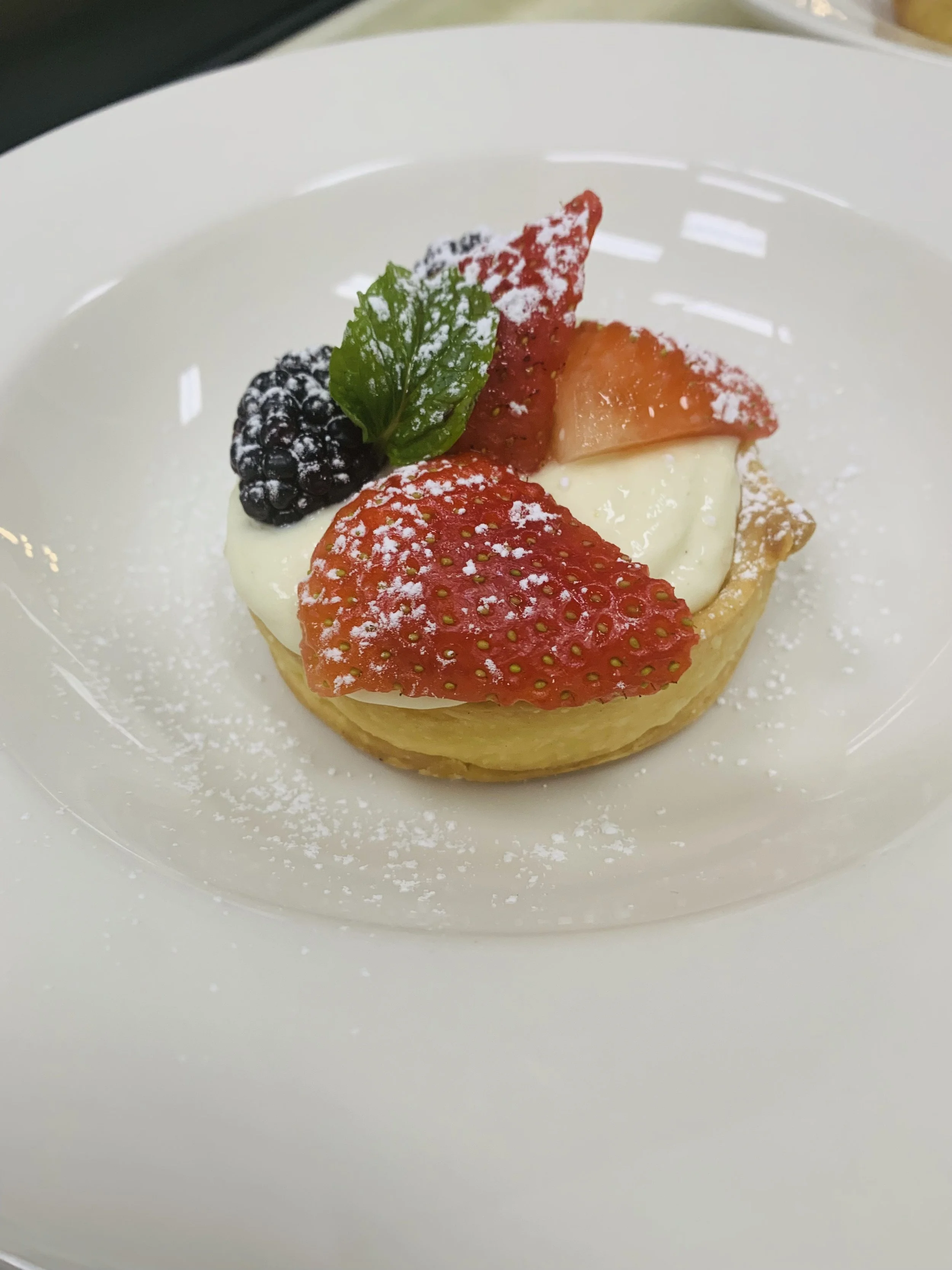 A dessert with strawberries, blackberries, and a green mint leaf on a crust, dusted with powdered sugar on a white plate.