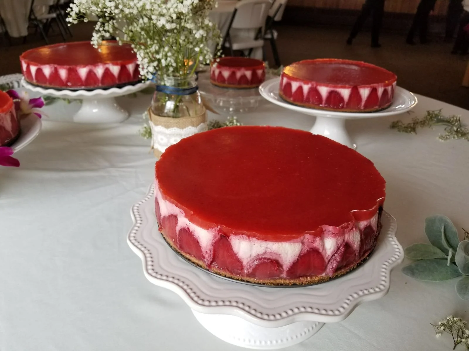 A table with several strawberry cheesecakes on white dessert stands, garnished with a small bouquet of white flowers in a jar and green leaves around the table.