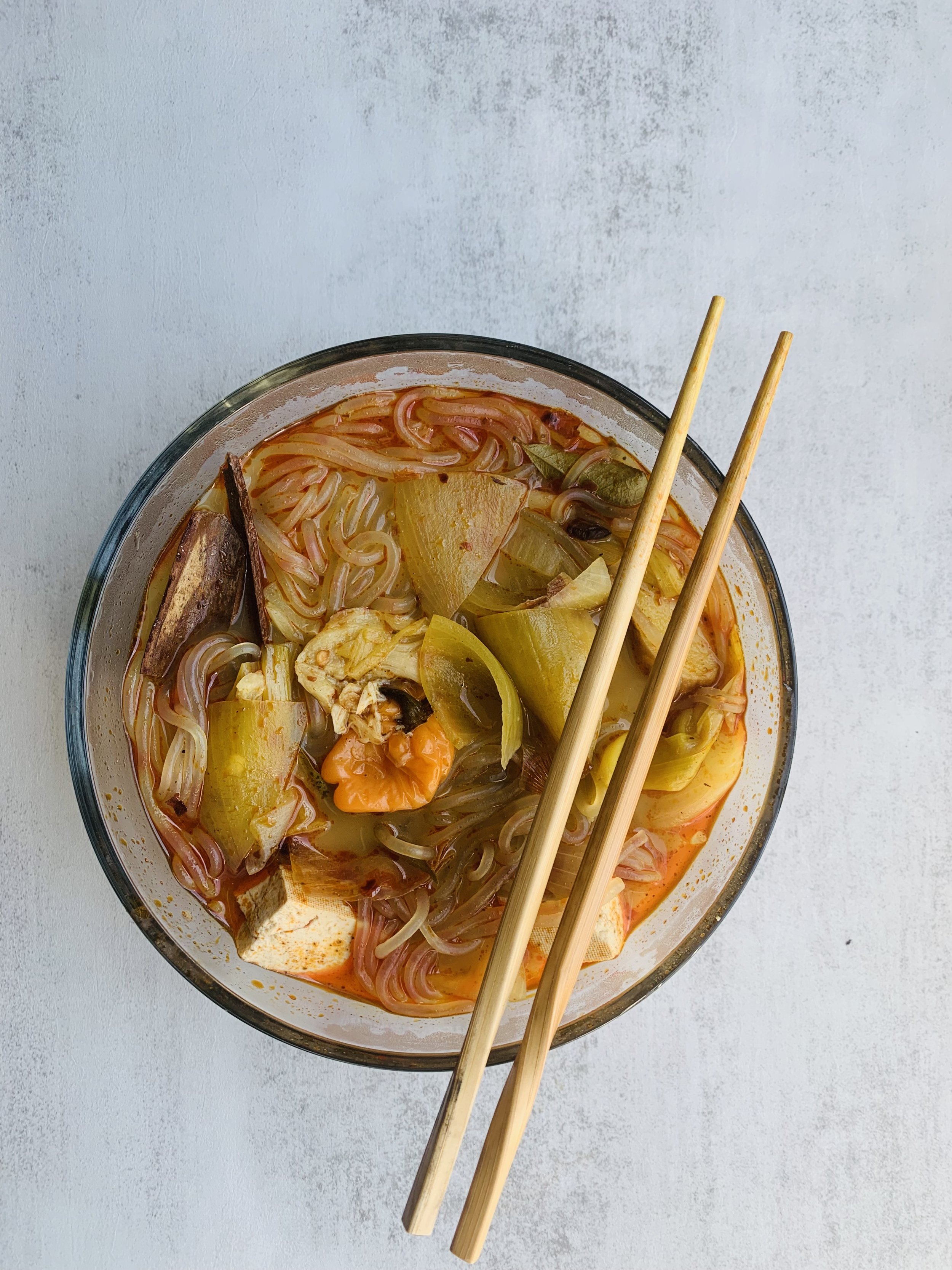 A bowl of noodle soup with vegetables, tofu, and chopsticks on a light gray surface.
