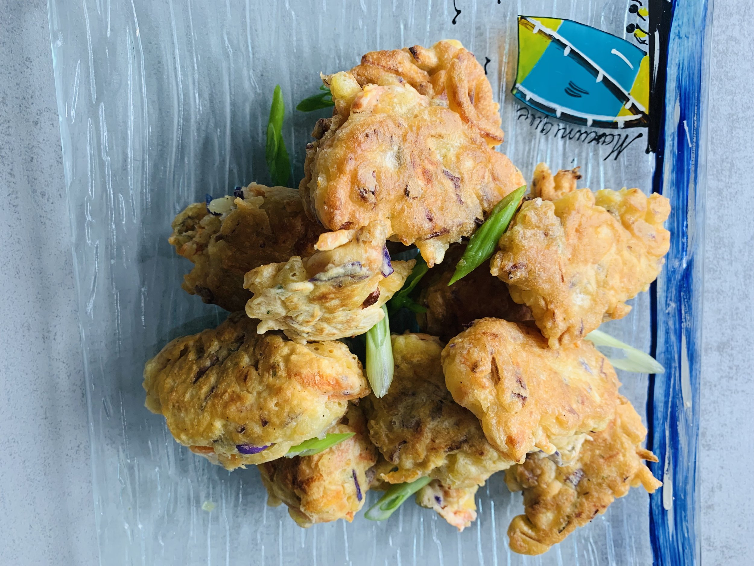 Assorted fried tempura vegetables and seafood served on a glass plate with a packet of sauce in the background.