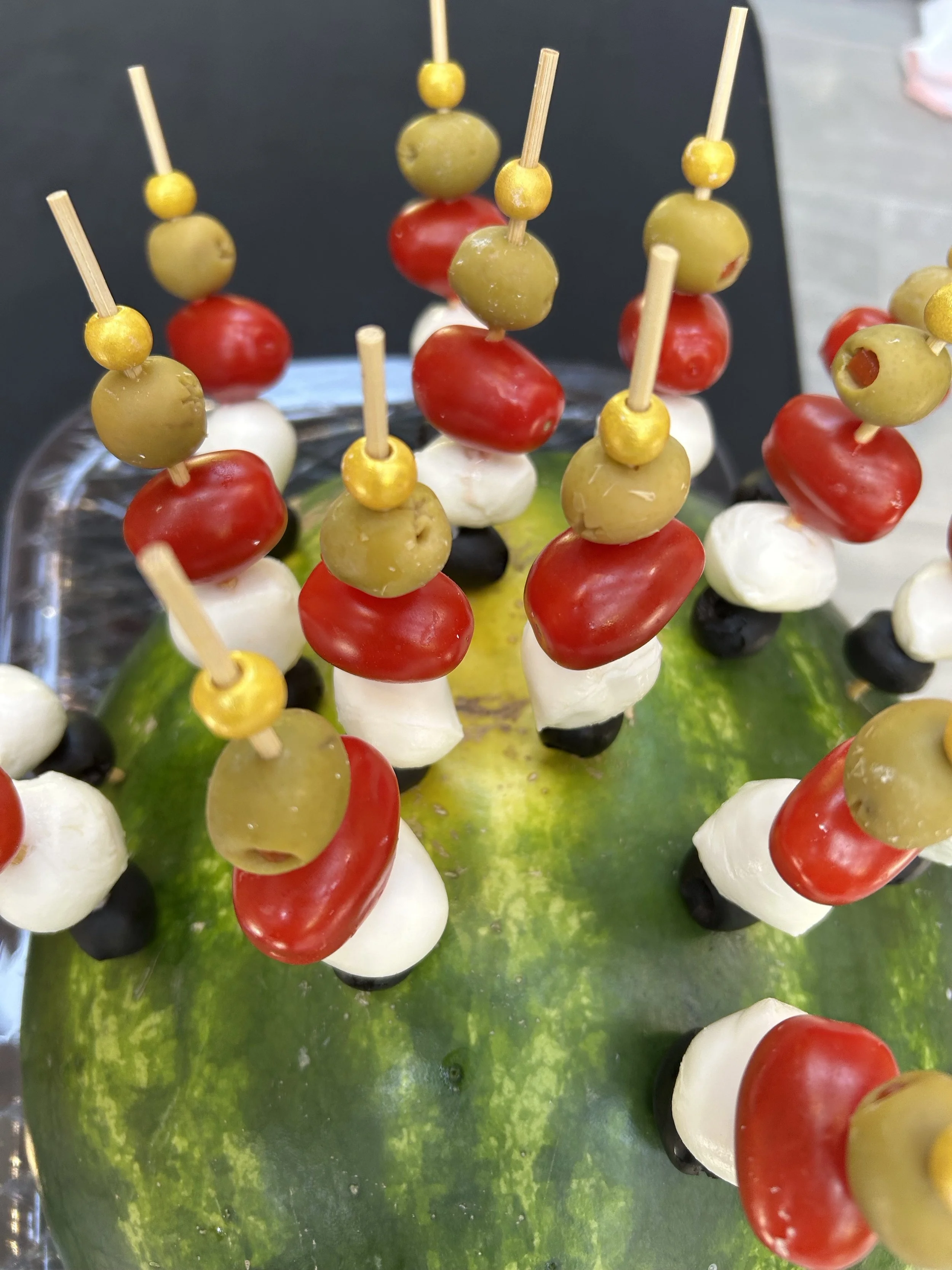 Skewers with green olives, cherry tomatoes, mozzarella balls, and black olives, arranged on a green watermelon.