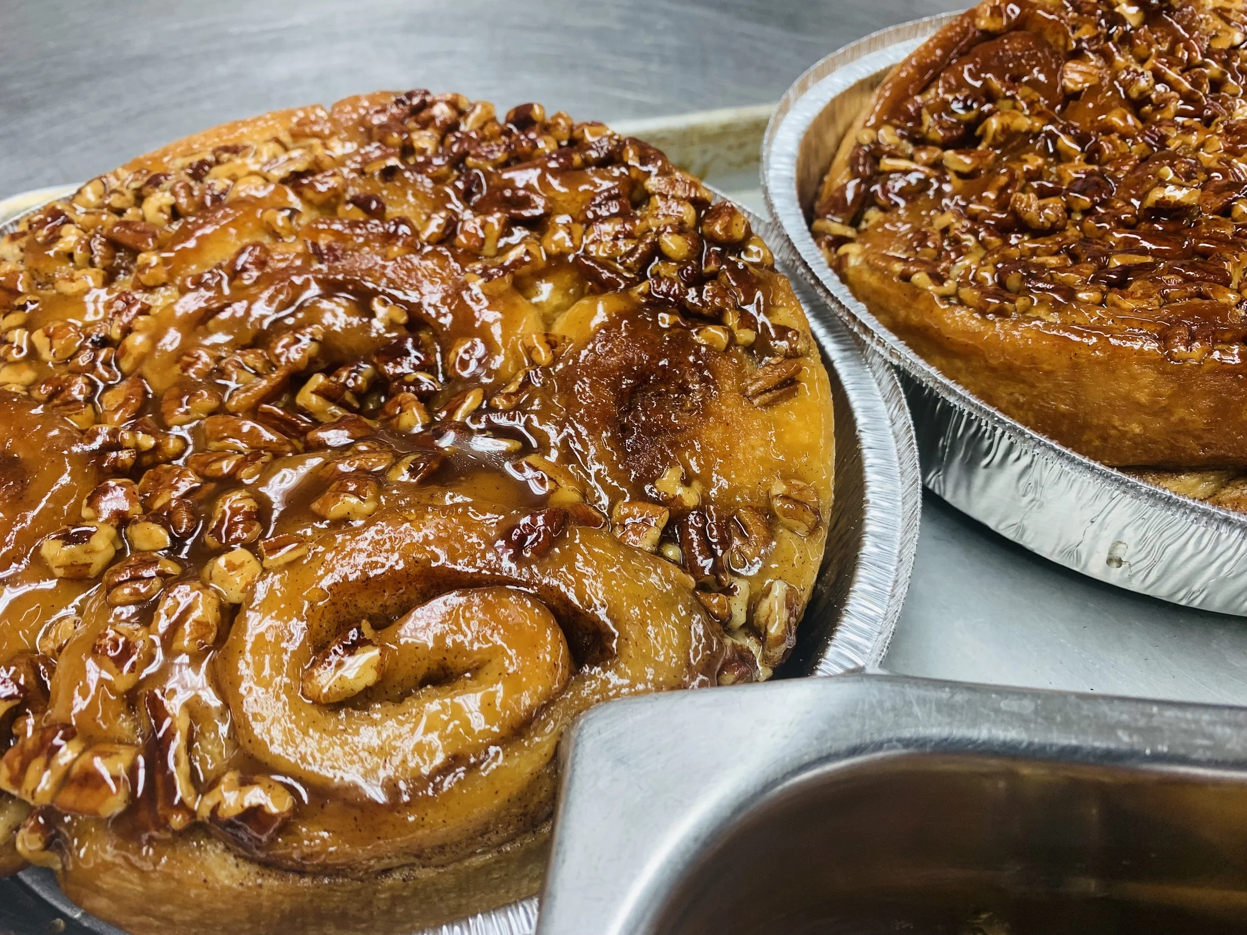 Two pecan pies in aluminum pans with caramel glaze on top, sitting on a stainless steel surface.