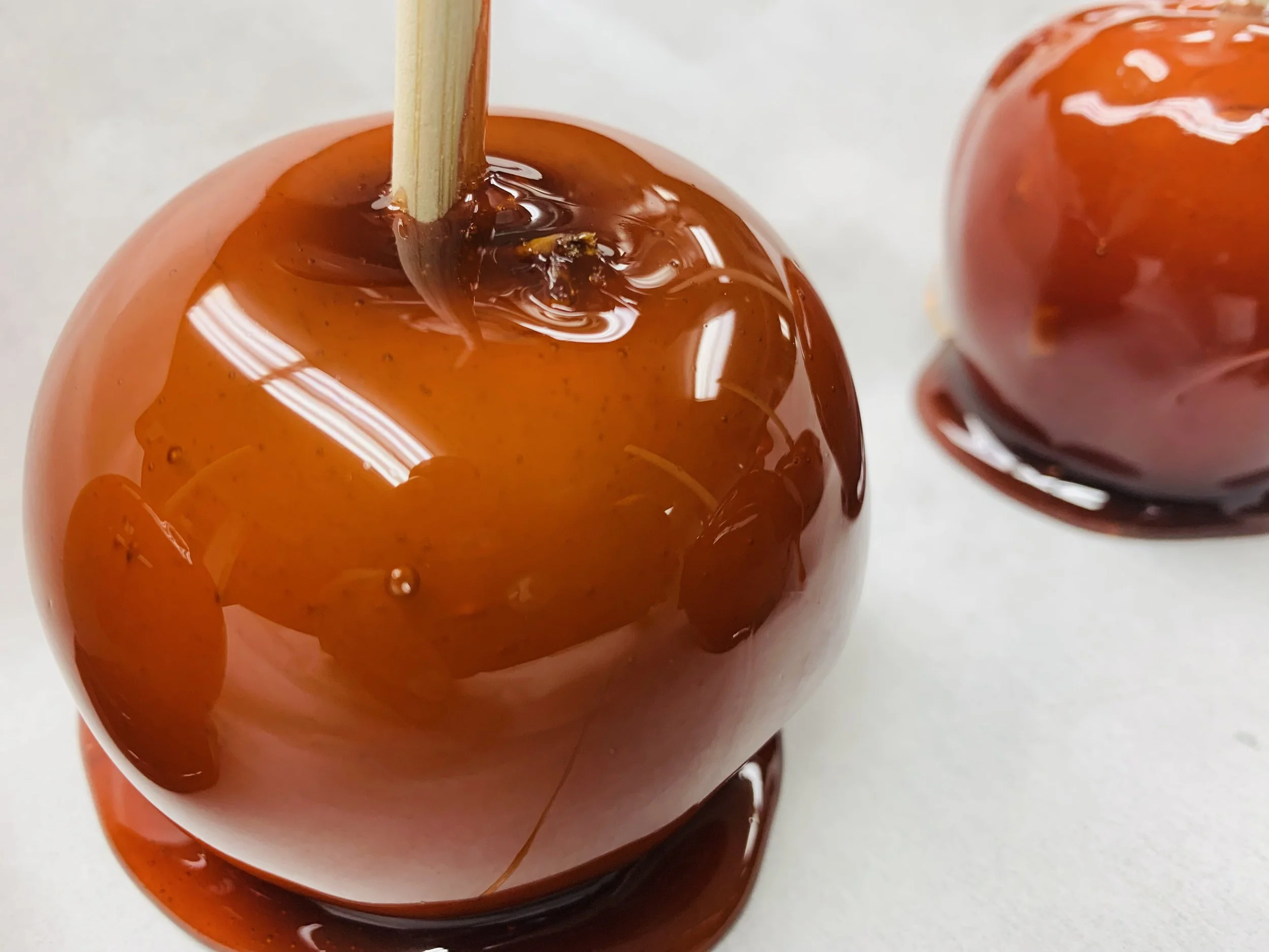 Close-up of two caramel apples on a white surface, one in the foreground with a wooden stick in it and the other in the background, both covered in shiny caramel.