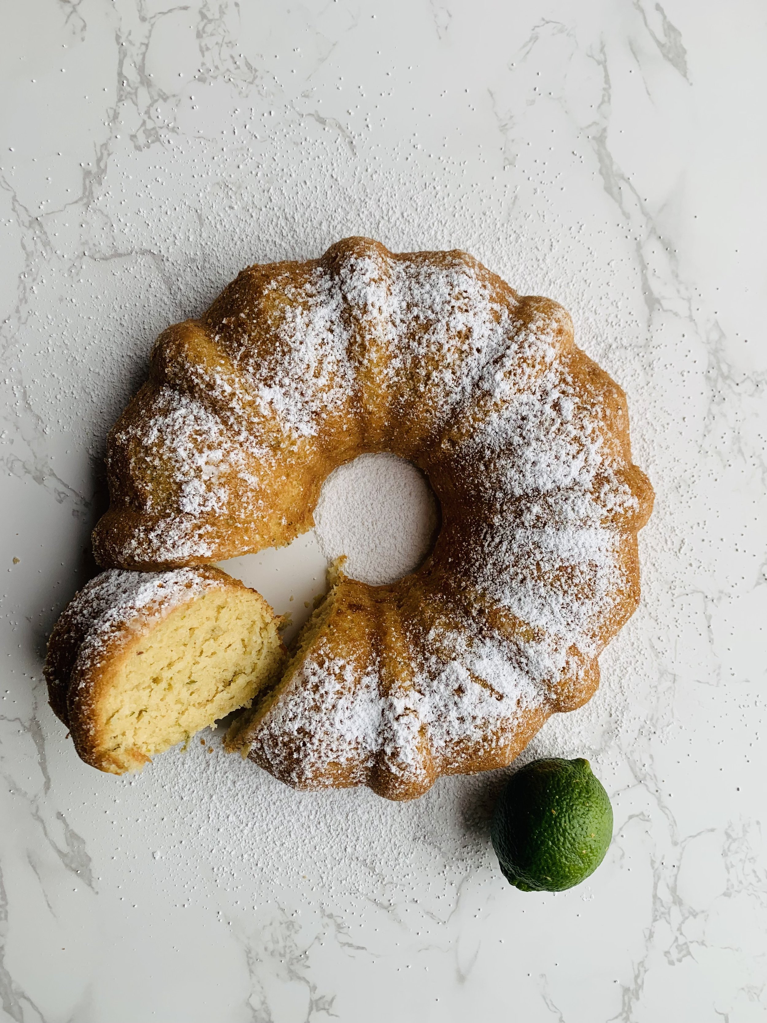 Bundt cake dusted with powdered sugar on a white marble surface, with a slice cut out revealing the inside, two limes, and scattered powdered sugar.