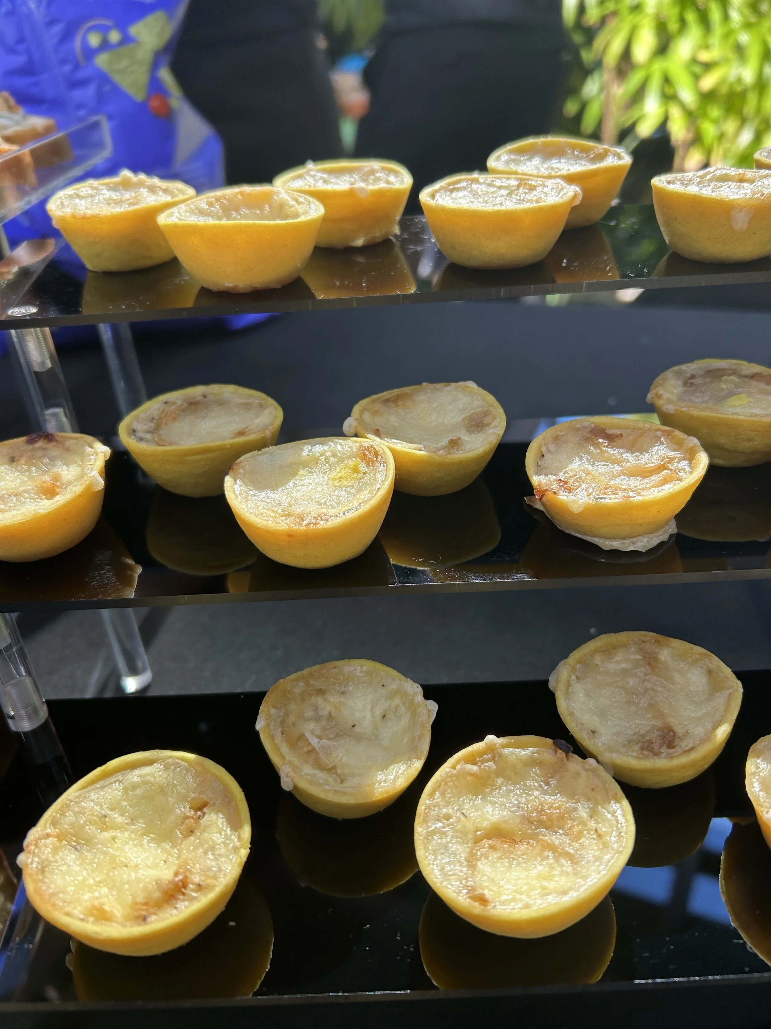 Lemon halves on a reflective black tray, some with grilled or roasted surfaces, outdoors with green foliage in background.