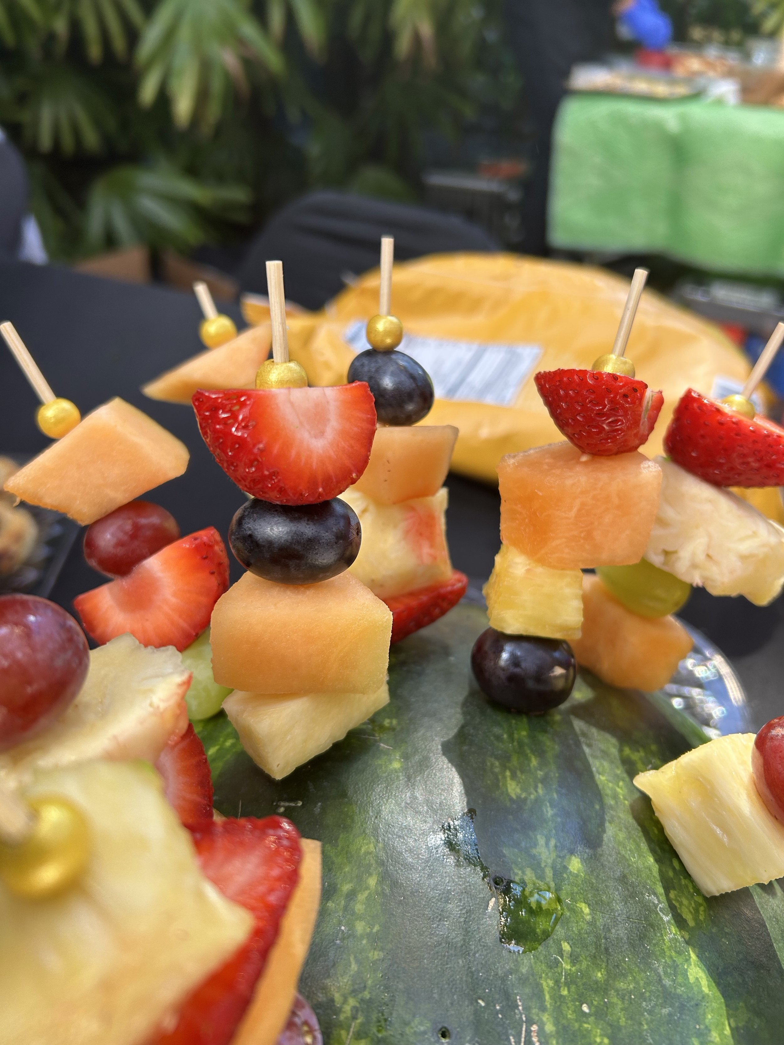 Close-up of assorted fruit skewers with strawberries, black grapes, cantaloupe, pineapple, and honeydew on a platter, with a blurred background of greenery and packaged sliced fruit.