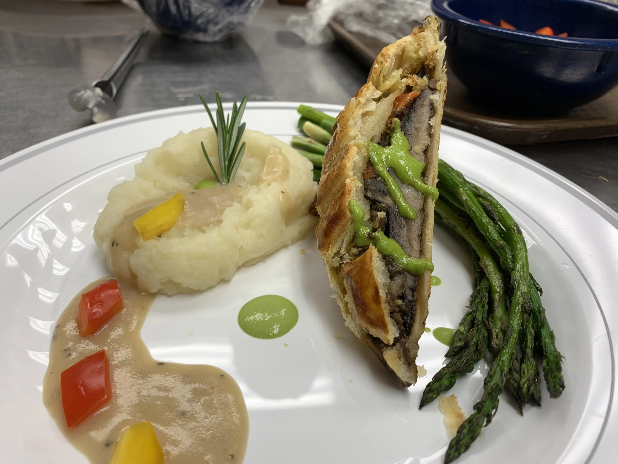 A plate with mashed potatoes topped with sprigs of rosemary, a serving of gravy with chopped red and yellow bell peppers, and a slice of vegetable pot pie with a golden crust, green beans on the side, and a small sprig of herbs on top.