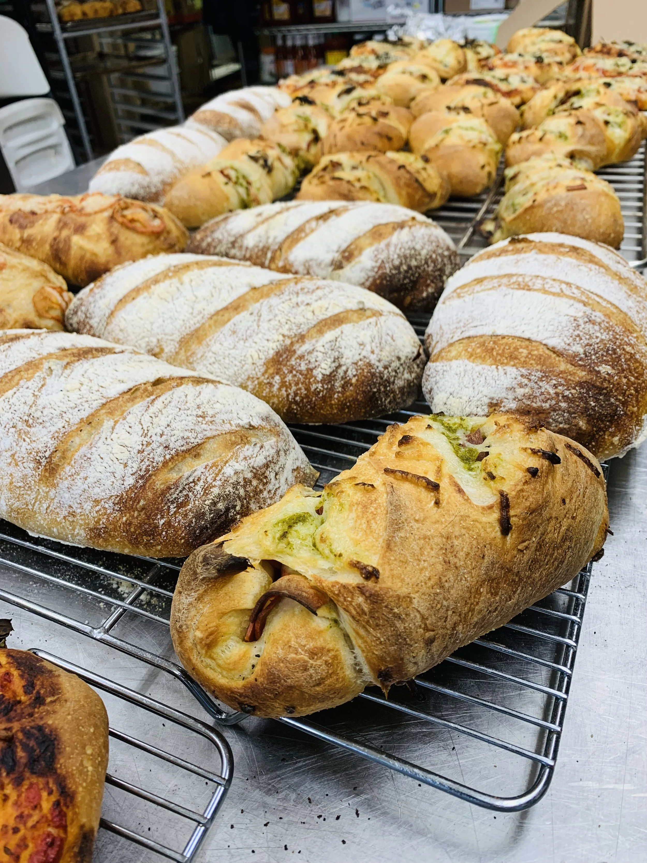 Assorted baked bread and pastries on cooling racks in a bakery, including round loaves dusted with flour and stuffed bread filled with vegetables and meats.