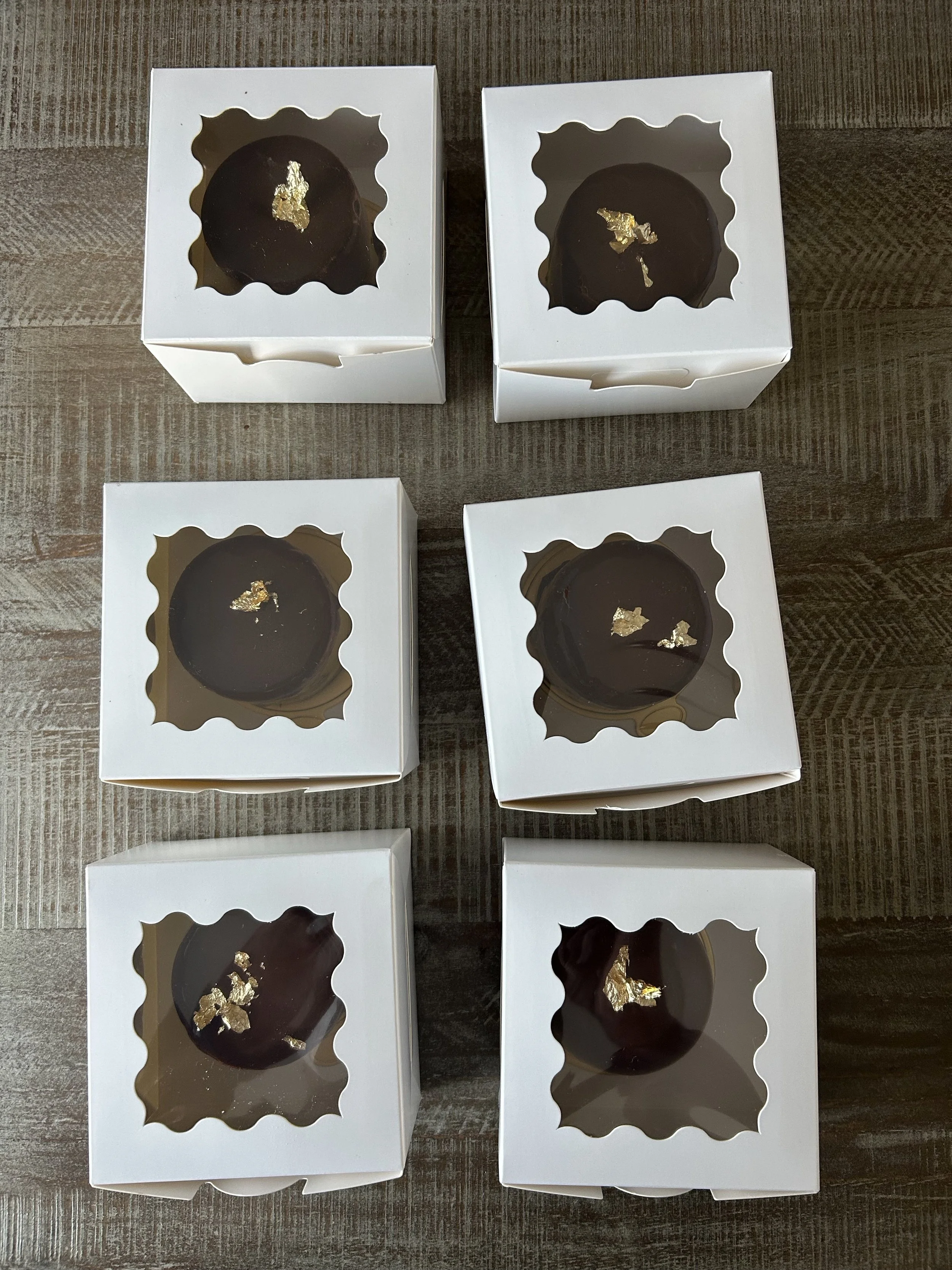 Six white boxes with transparent windows showing dark chocolates topped with gold leaf on a wooden surface.