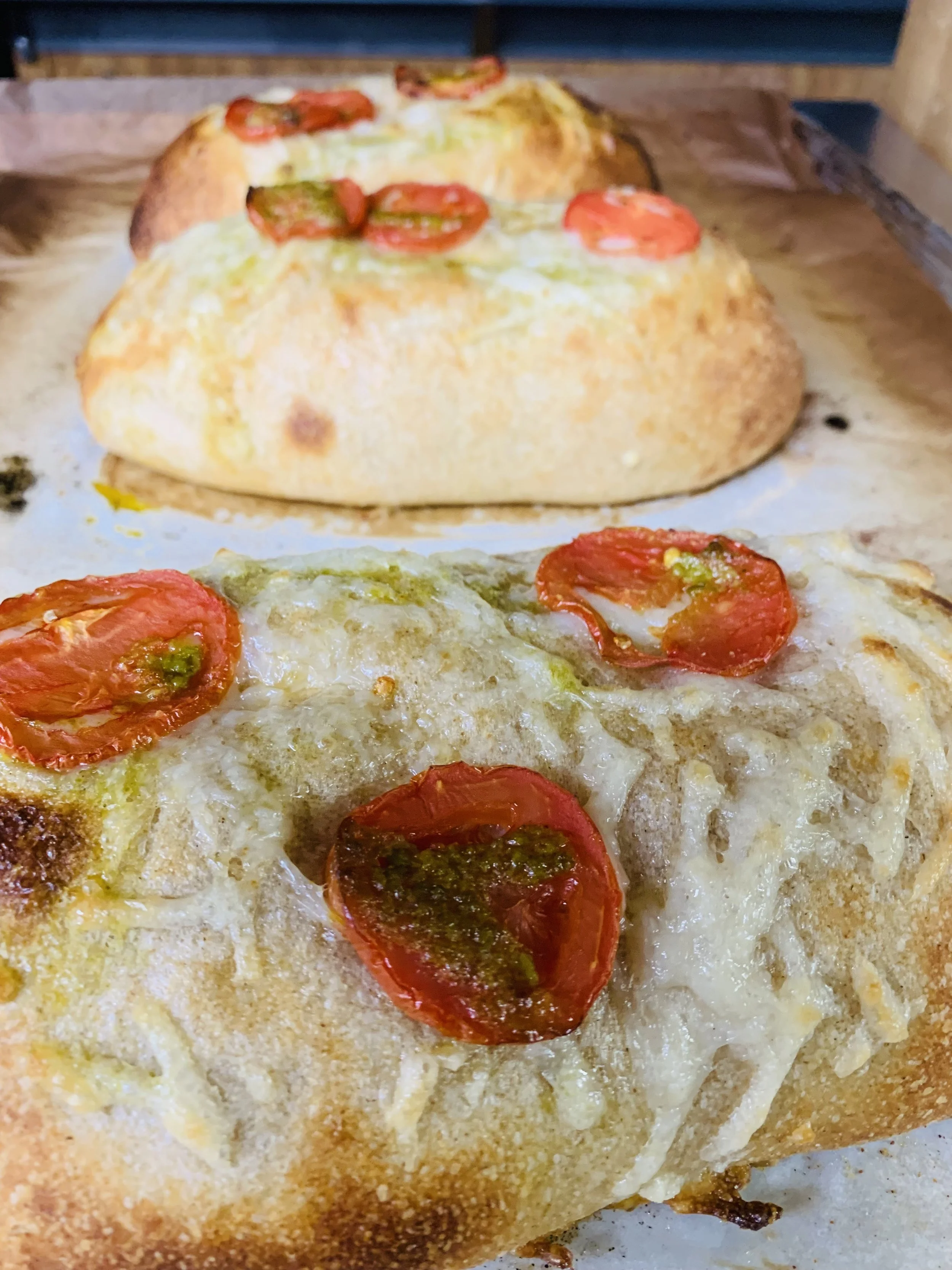 Close-up of a pizza with melted cheese, cherry tomato slices, and green pesto on top, on a baking sheet.