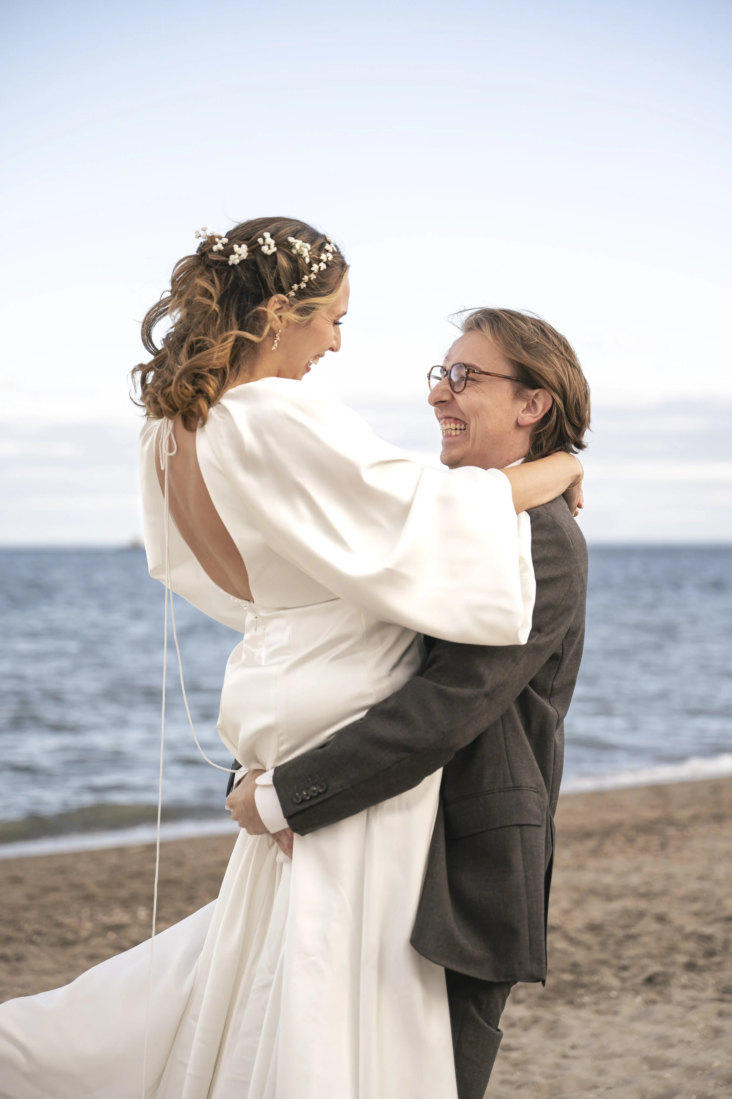 Close-up of a bride and groom's hands showing wedding rings, with the bride wearing a ring with a heart-shaped stone, and the groom wearing a plain band. The bride's hand with rings rests on the groom's hand, which is adorned with a watch, as they hold hands.