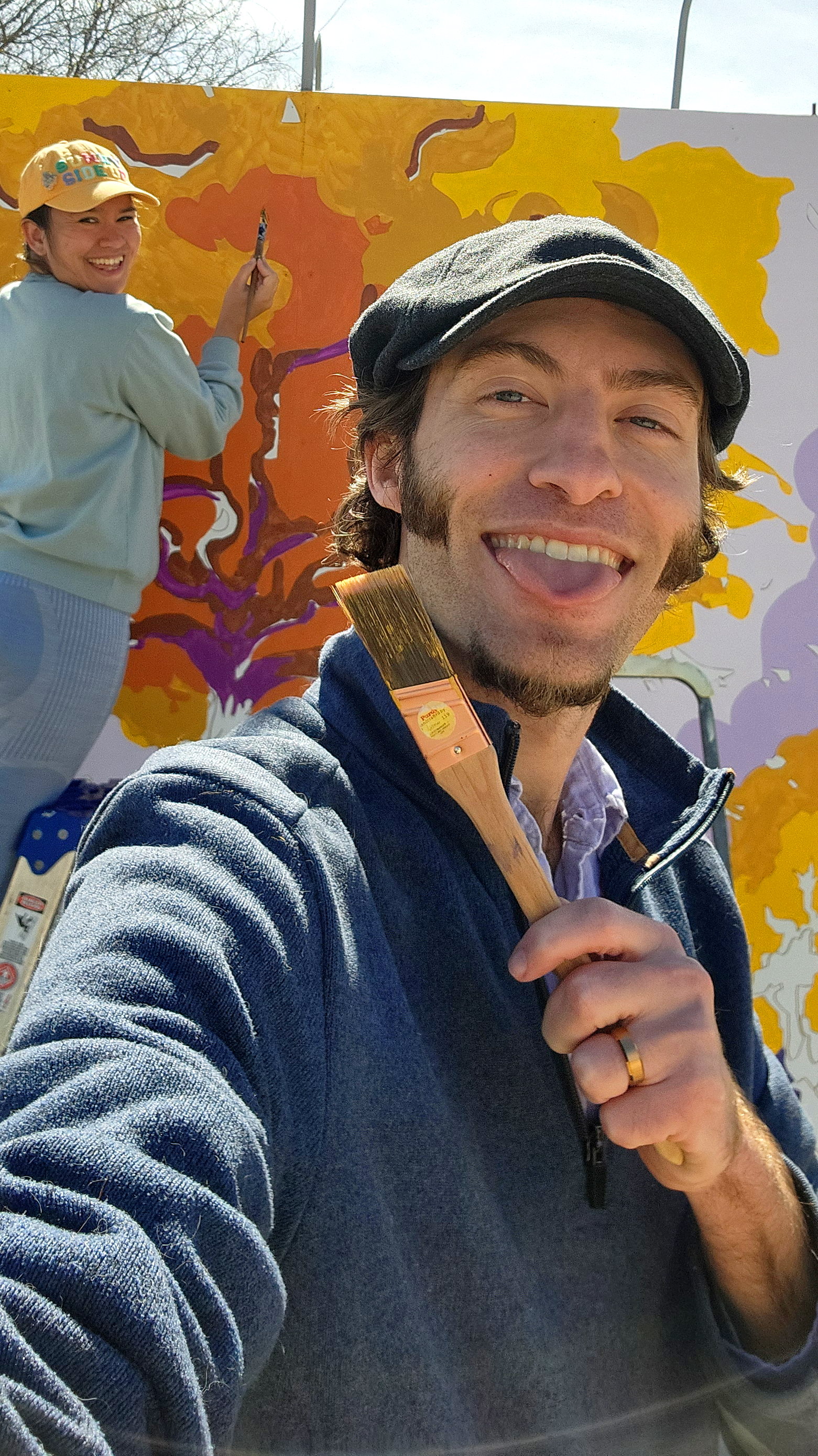 Two people painting a large outdoor mural featuring a tree with yellow and orange leaves. The man in the foreground is smiling and holding a paintbrush, while the woman in the background is painting on the mural.