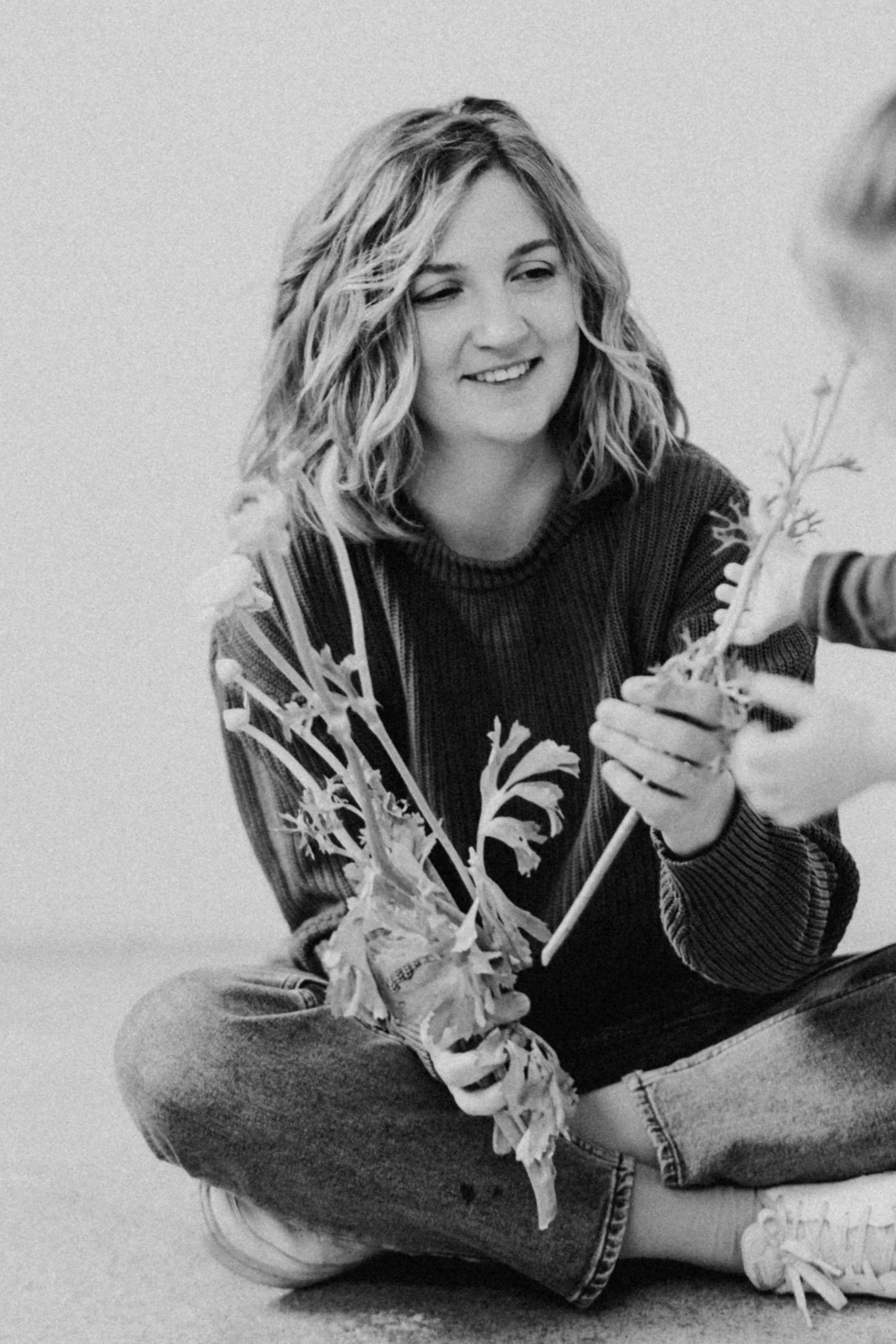 Schwarz-weiße Fotografie einer lachenden jungen Frau mit lockigem, schulterlangem Haar, die im Sitzen auf dem Boden sitzt. Sie hält Ranunkeln in der Hand und reicht diese einem Kind, das man nur schemenhaft erkennt.