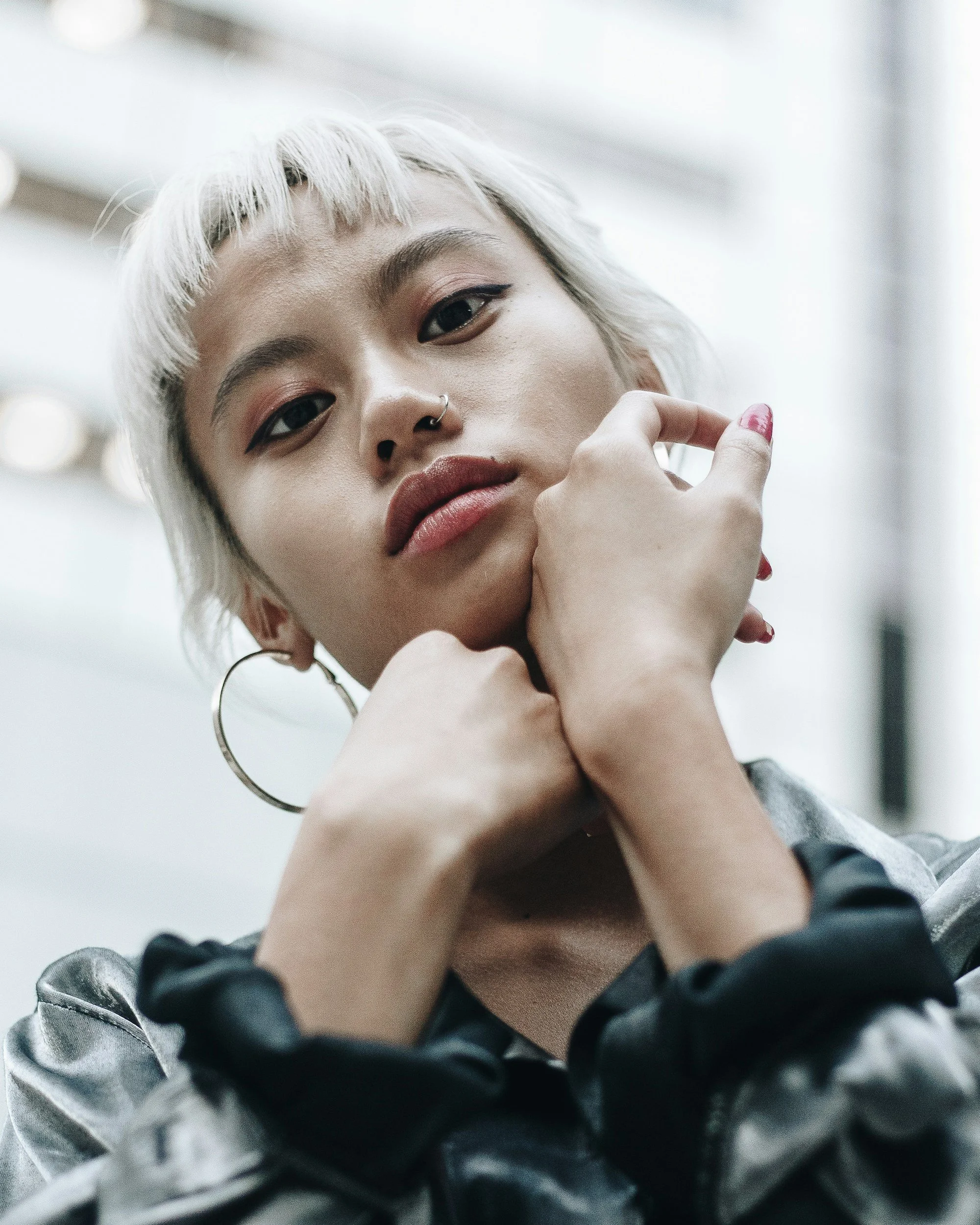 Close-up of a person with short white hair, wearing hoop earrings, a nose ring, and a metallic jacket, looking into the camera with one hand on their chin.