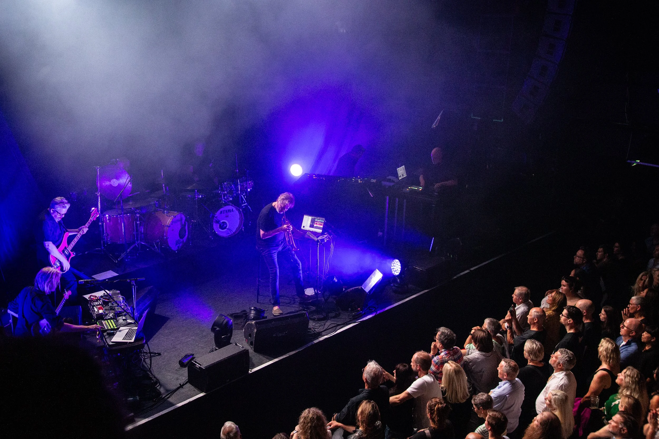 A live music concert with a band performing on stage illuminated by purple lights, and a seated audience watching.