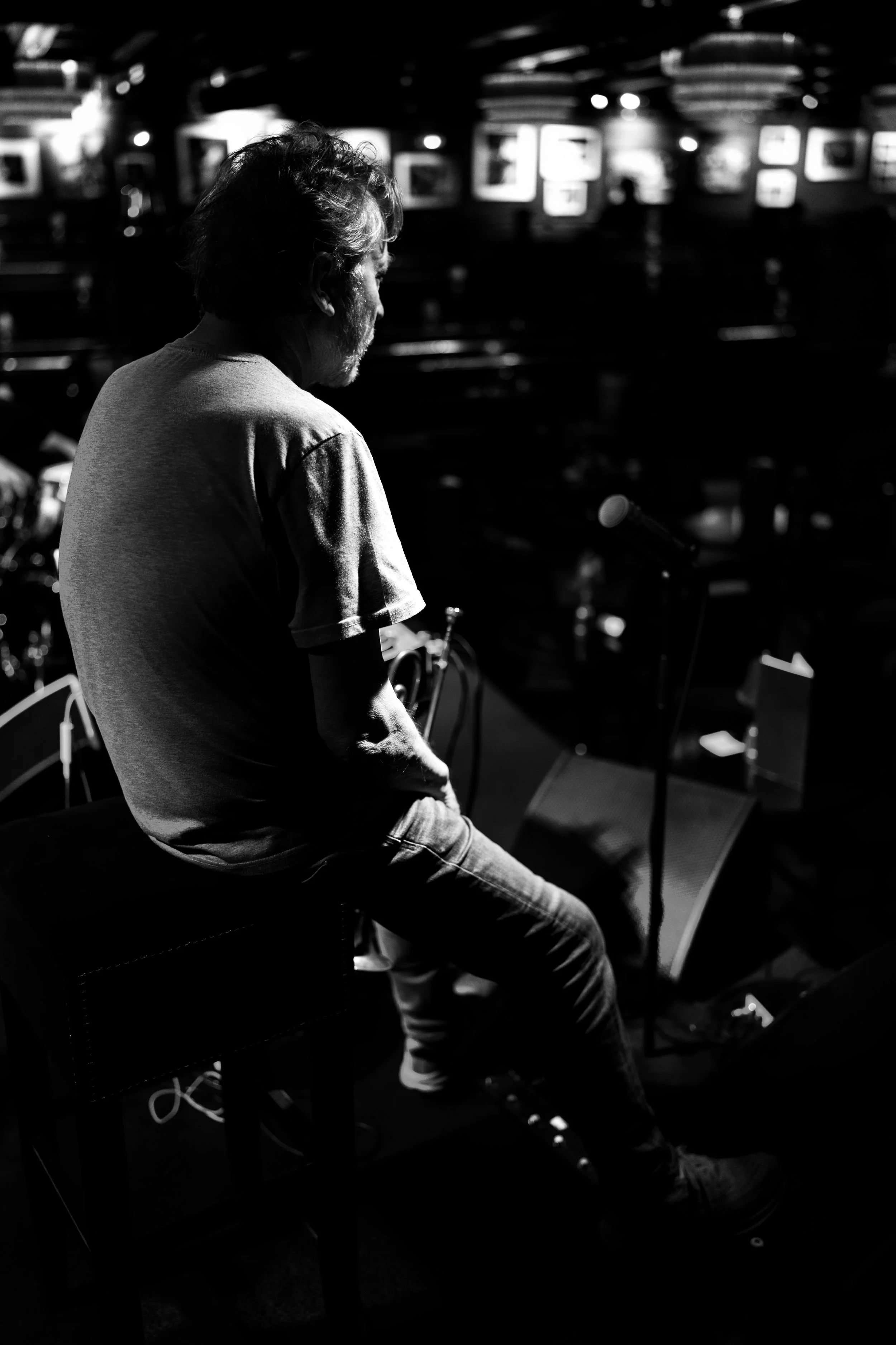 A black and white photo of a man sitting on a stool on stage with a microphone, in a dimly lit venue.