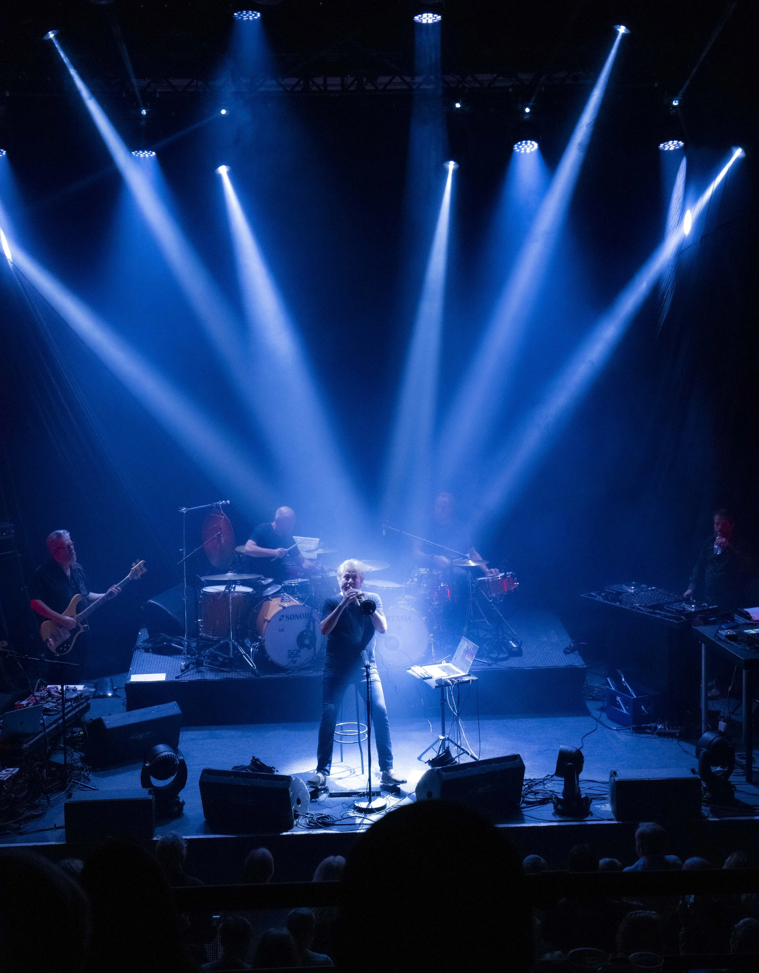 Nils Petter Molvaer 's Khmer performing on stage with blue stage lights and fog, with a singer at center and other band members playing instruments.
