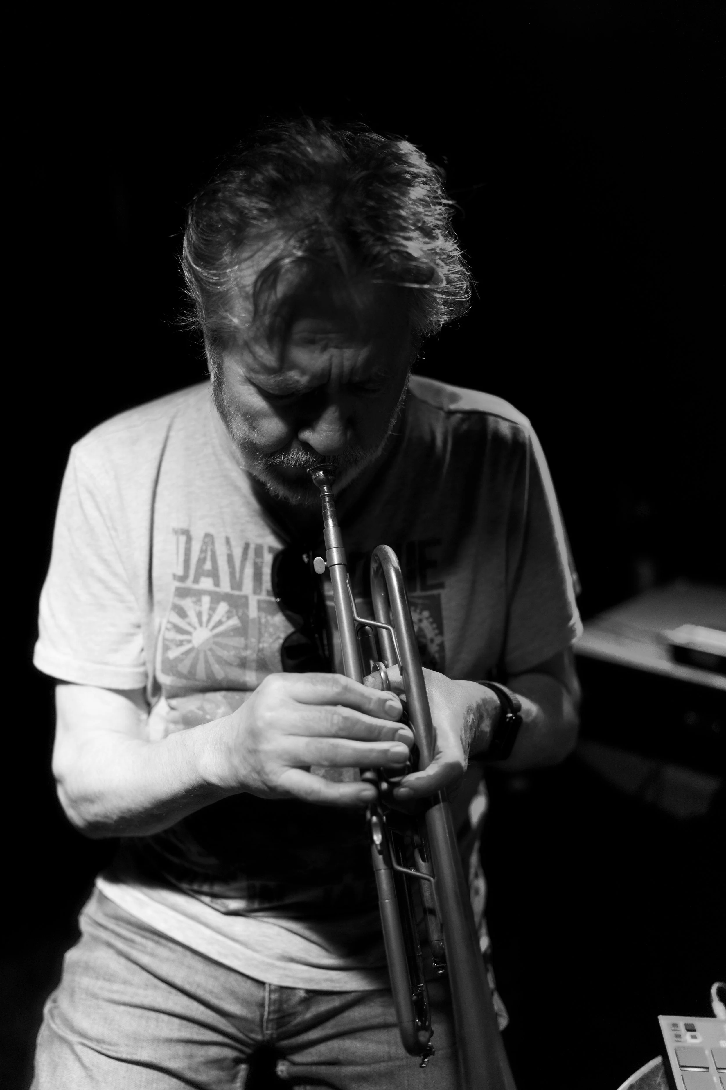 A man playing a trumpet in a dark room, wearing a light-colored T-shirt, black wristwatch, and sunglasses hanging from his shirt collar.