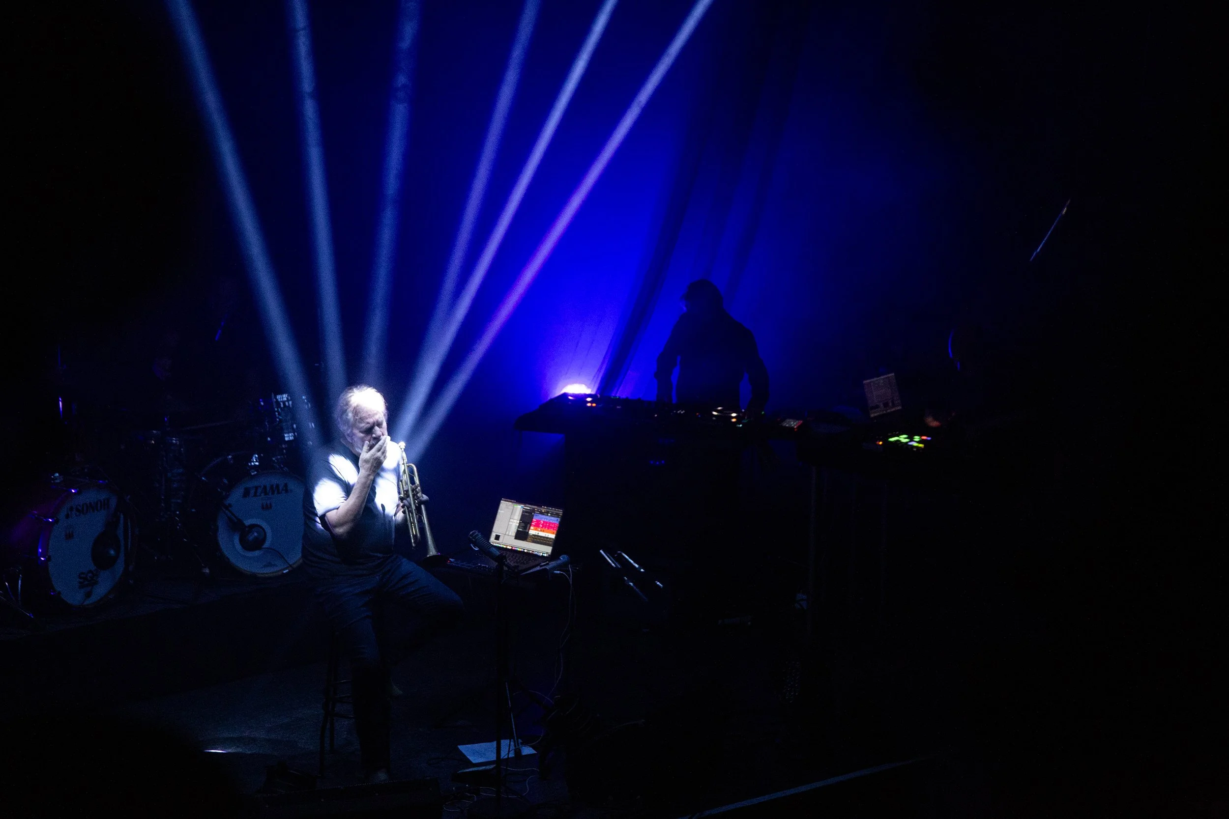 A musician with gray hair playing a trumpet on stage, illuminated by blue stage lights, with DJ equipment and drums in the background.