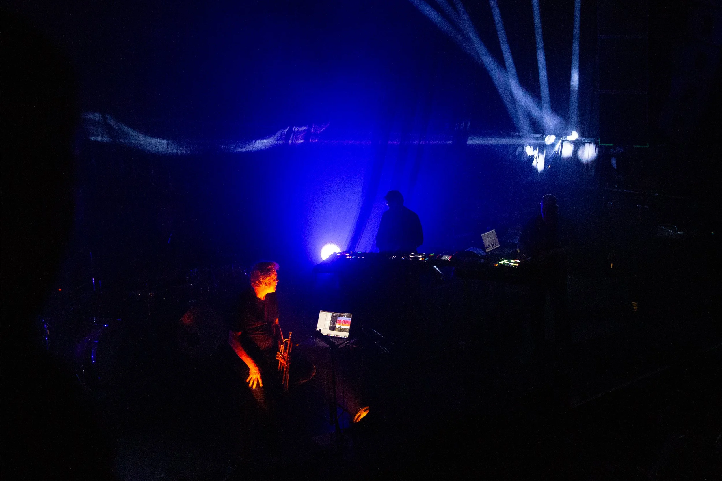 Silhouettes of musicians performing on stage with blue and purple lighting, electronic equipment, and a dark background.