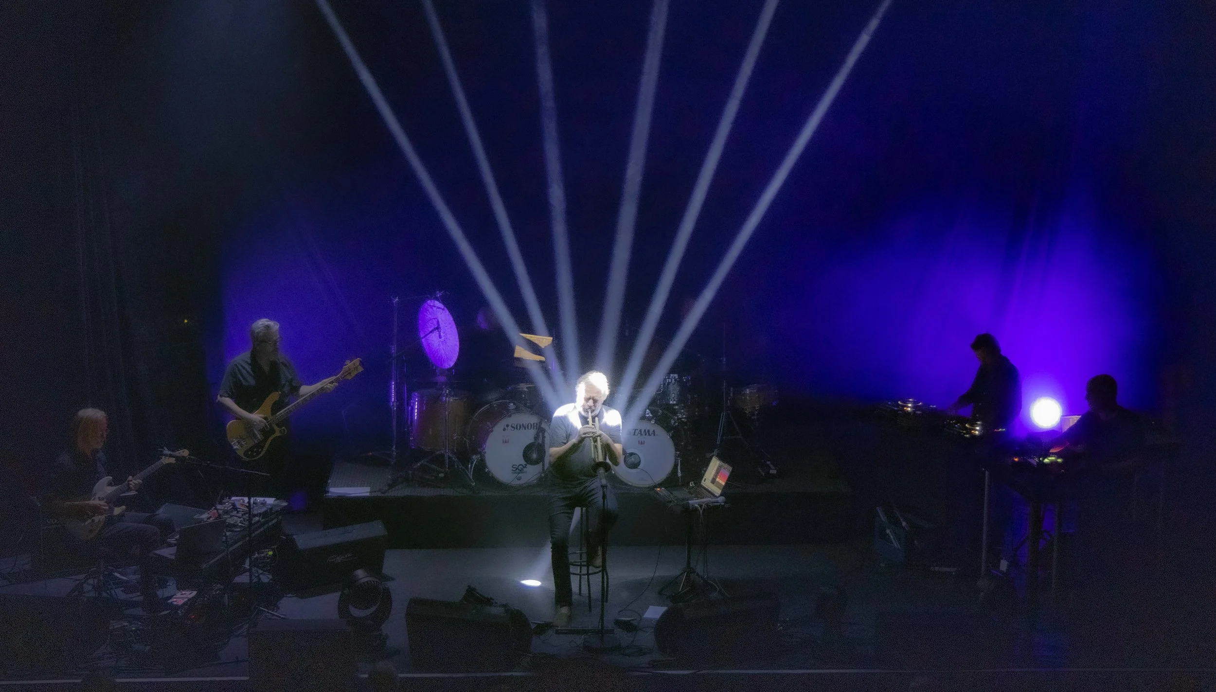 Nils Petter Molvaer 's Khmer on stage with purple and blue lighting, a drummer behind a drum set, a bassist left of center, a guitarist on the far left, and two keyboard players on the far right, with bright stage lights shining behind them.