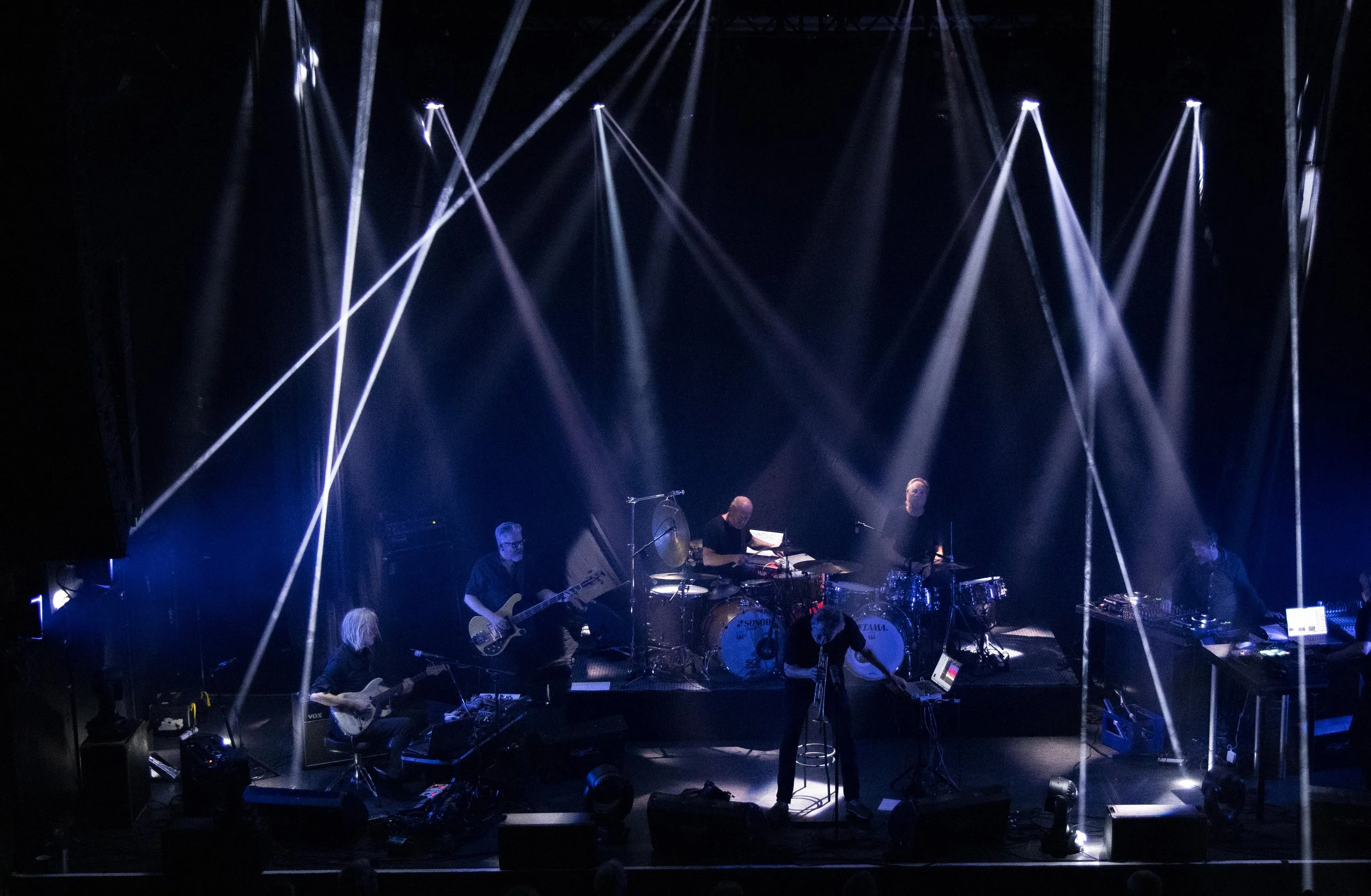 Nils Petter Molvaer 's Khmer performs on stage with multiple musicians, instruments, and bright stage lighting with beams of white light overhead.