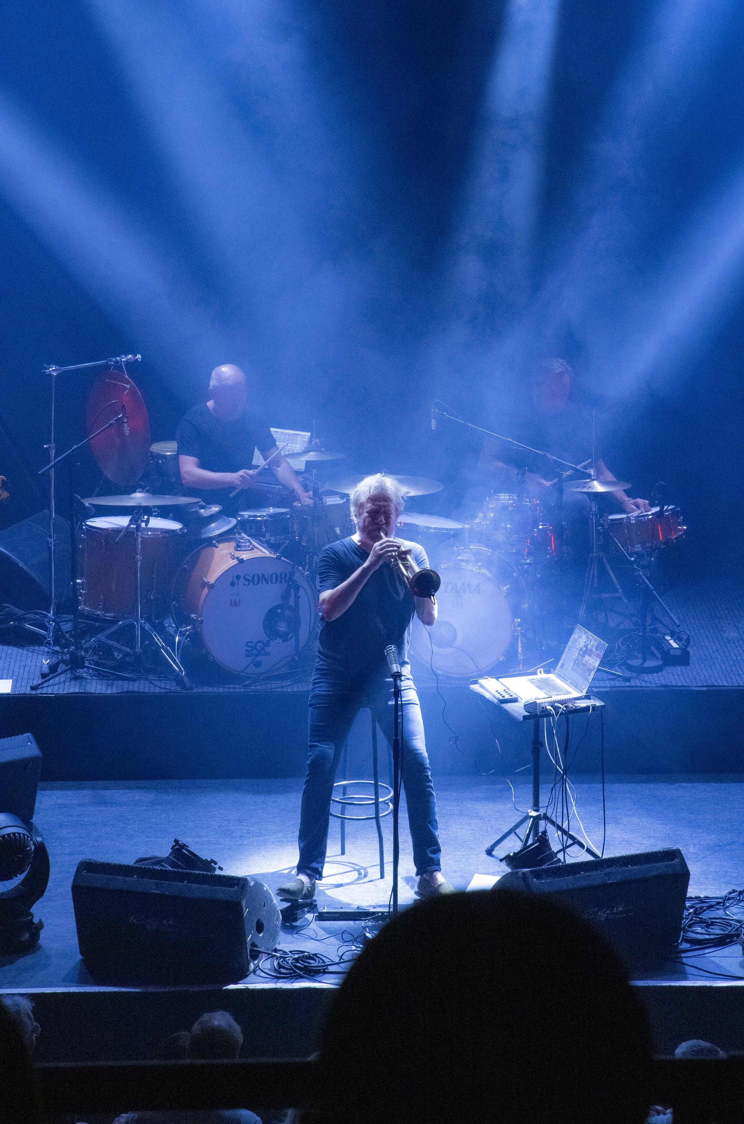 Nils Petter Molvaer on stage, playing into a microphone with a trumpet, with blue stage lights shining down on him. Two percussionists play drums in the background, and electronic equipment is set up nearby.