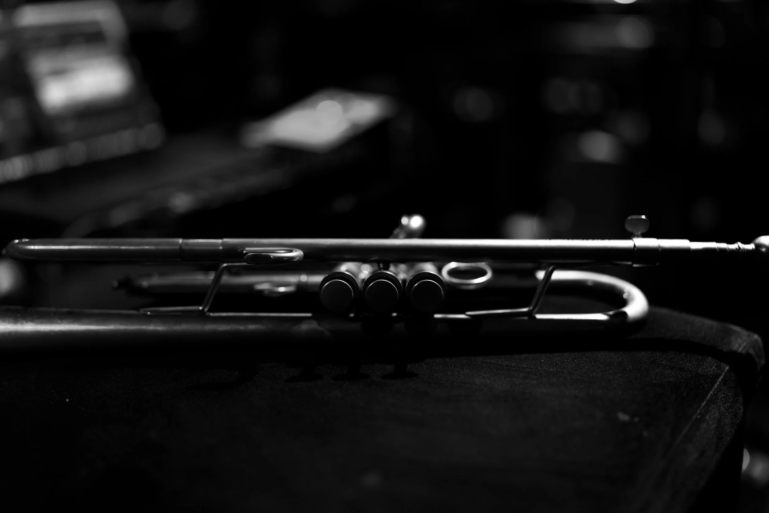 Black and white photo of a trumpet resting on a dark surface with blurred equipment in the background.